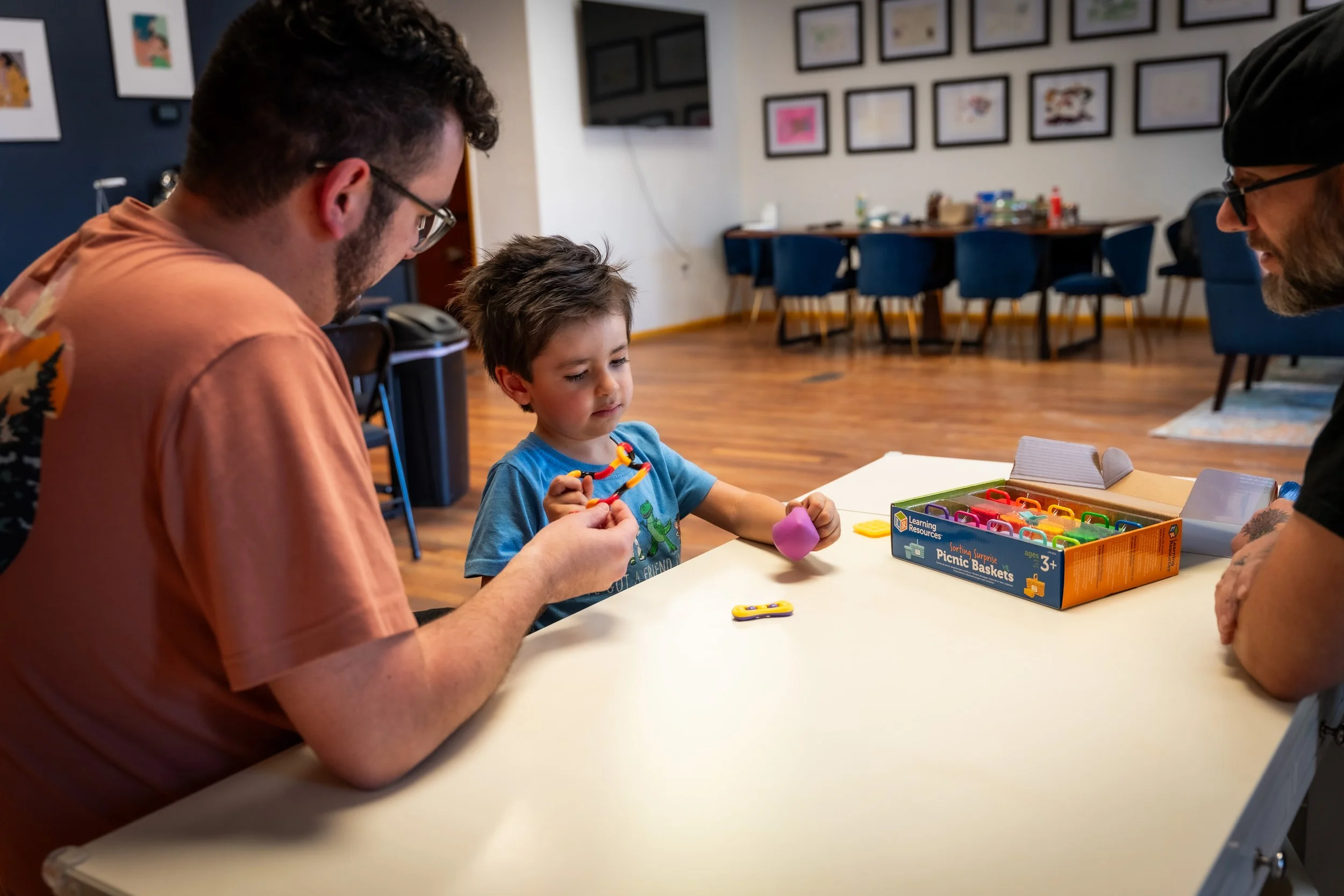 A young boy sitting at a table playing with colorful building toys, with two men, possibly family members, engaging with him. The background shows a wall decorated with framed artwork and a table with drinks and supplies.
