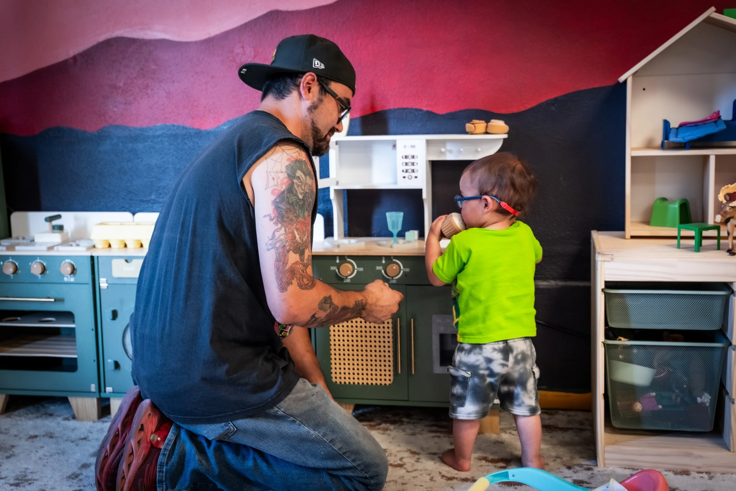 A man with tattoos on his arm wearing glasses and a black backwards cap kneeling beside a young boy with glasses, wearing a bright green shirt, in a playroom. The boy is drinking from a cup, and there are toys and furniture around them.