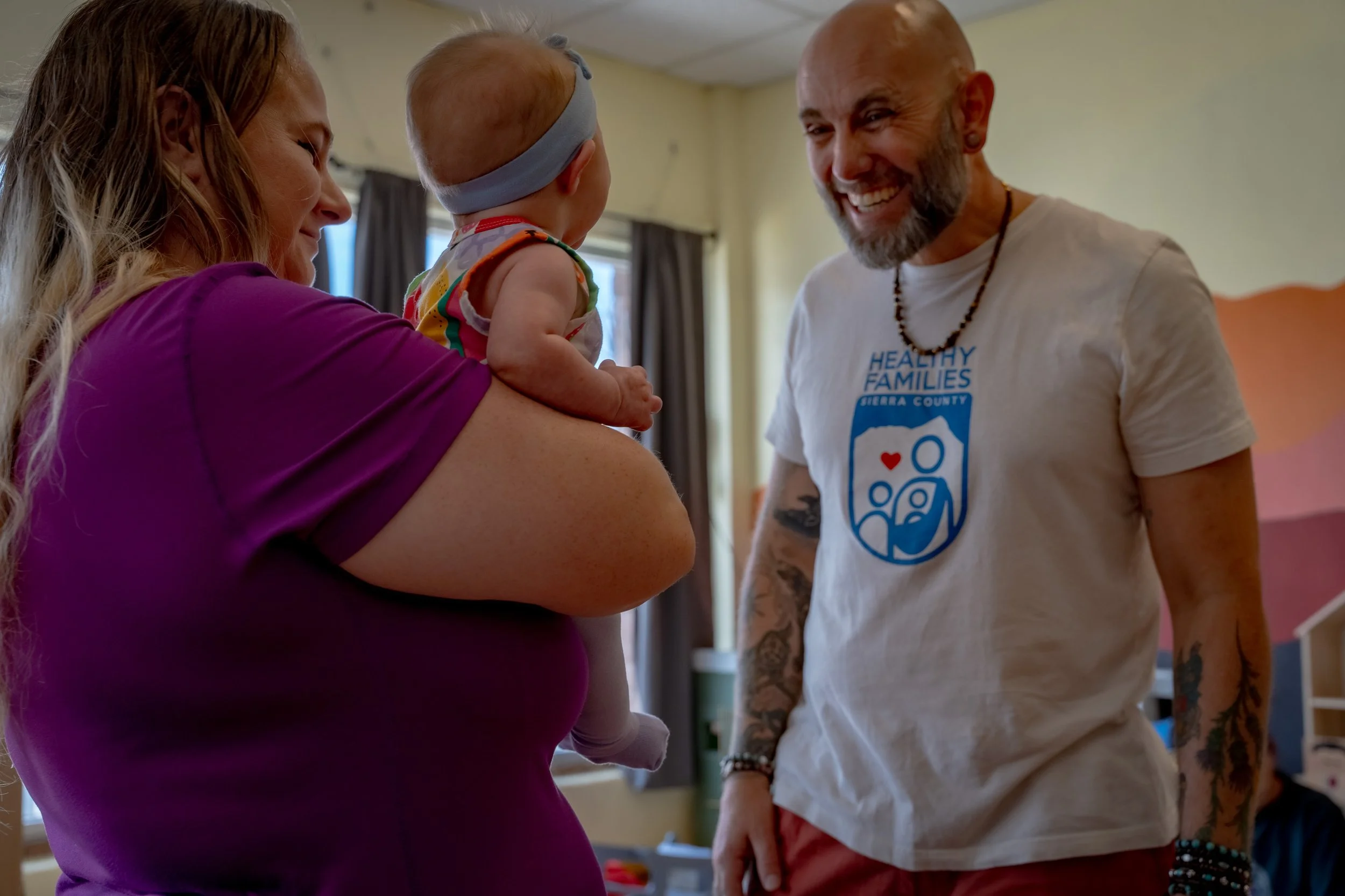 A woman is holding a baby with a headband, and a man with tattoos and a white t-shirt is standing nearby, smiling.