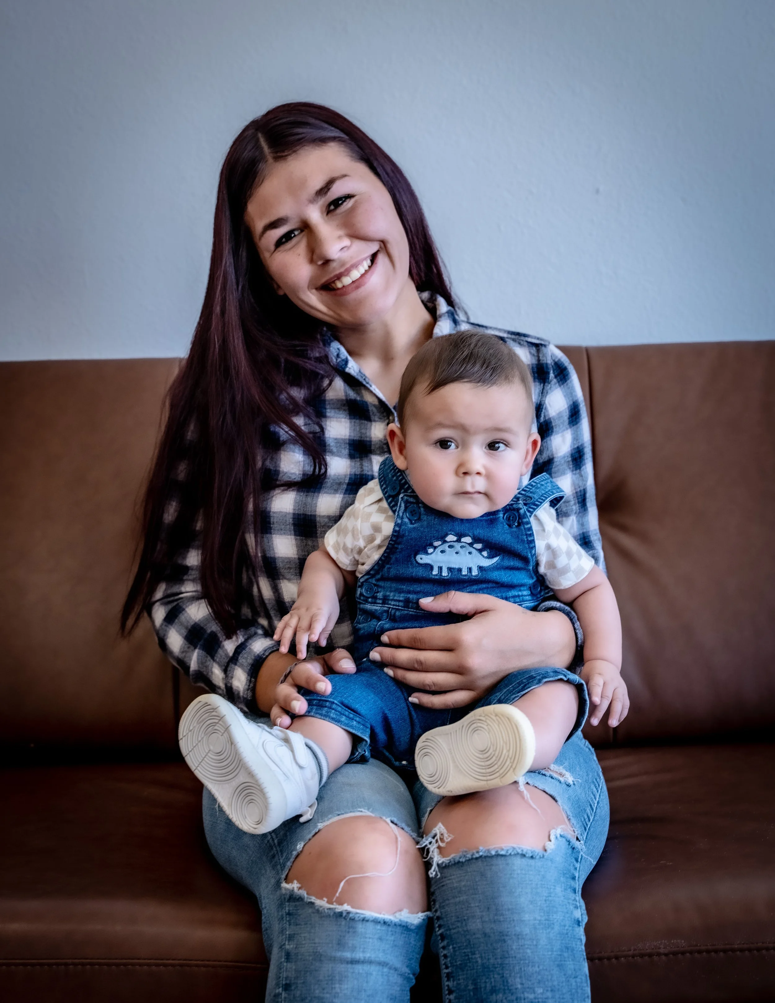 A smiling woman with long dark hair sitting on a brown leather couch, holding a young boy on her lap. The boy is wearing a denim outfit with a dinosaur patch and white shoes. The woman is wearing a plaid shirt and ripped jeans. They are in a room with a plain, light-colored wall.