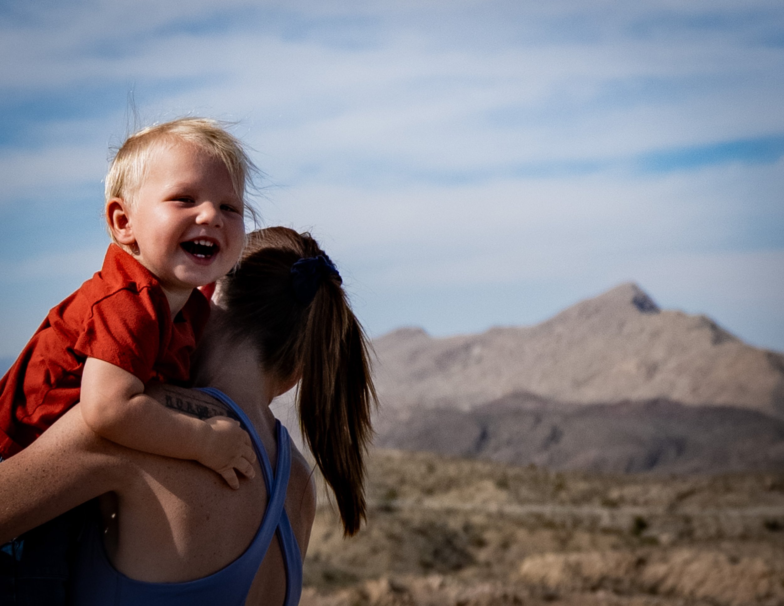 A young boy with blonde hair smiling and sitting on an adult's shoulders outdoors with a mountainous landscape and blue sky in the background.