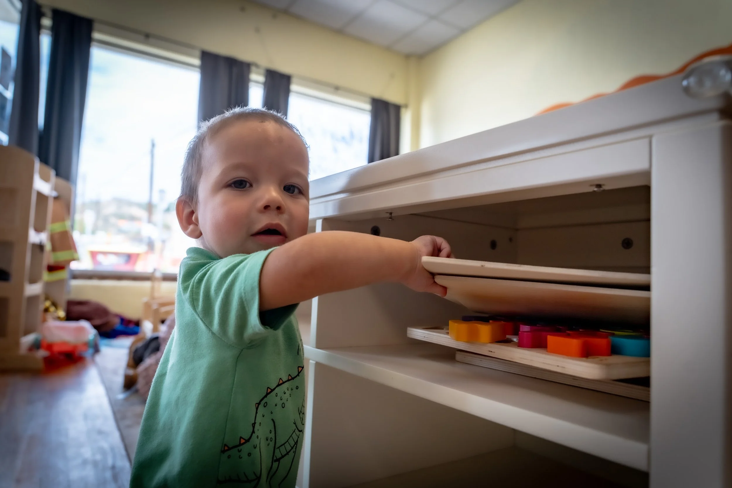 A young boy with light skin and short hair, wearing a green t-shirt with a dinosaur graphic, is reaching into a white shelf in a daycare or preschool setting. The background shows bright windows with blue curtains, and cluttered toys and furniture typical of a playroom.