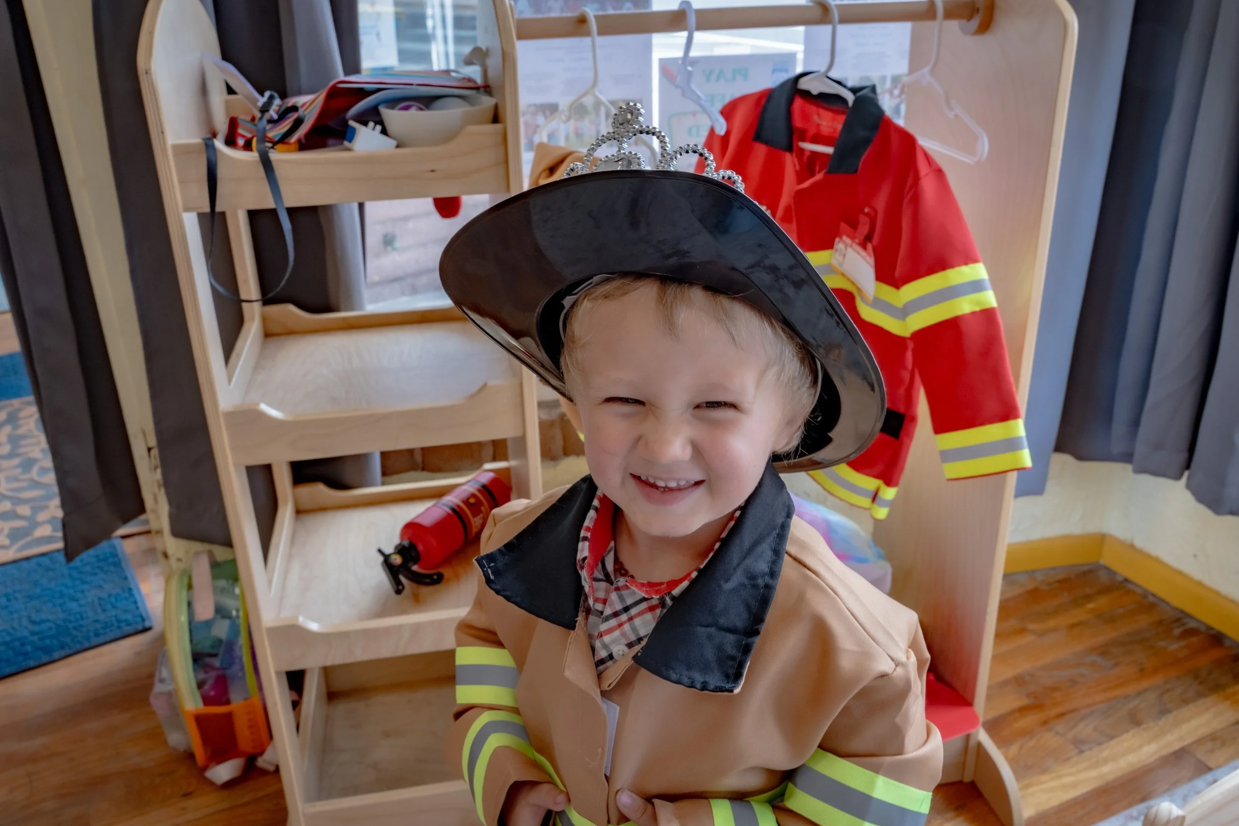 Child dressed as a firefighter with a black hat adorned with a small tiara, smiling in front of a wooden display with firefighter gear and coat hangers.