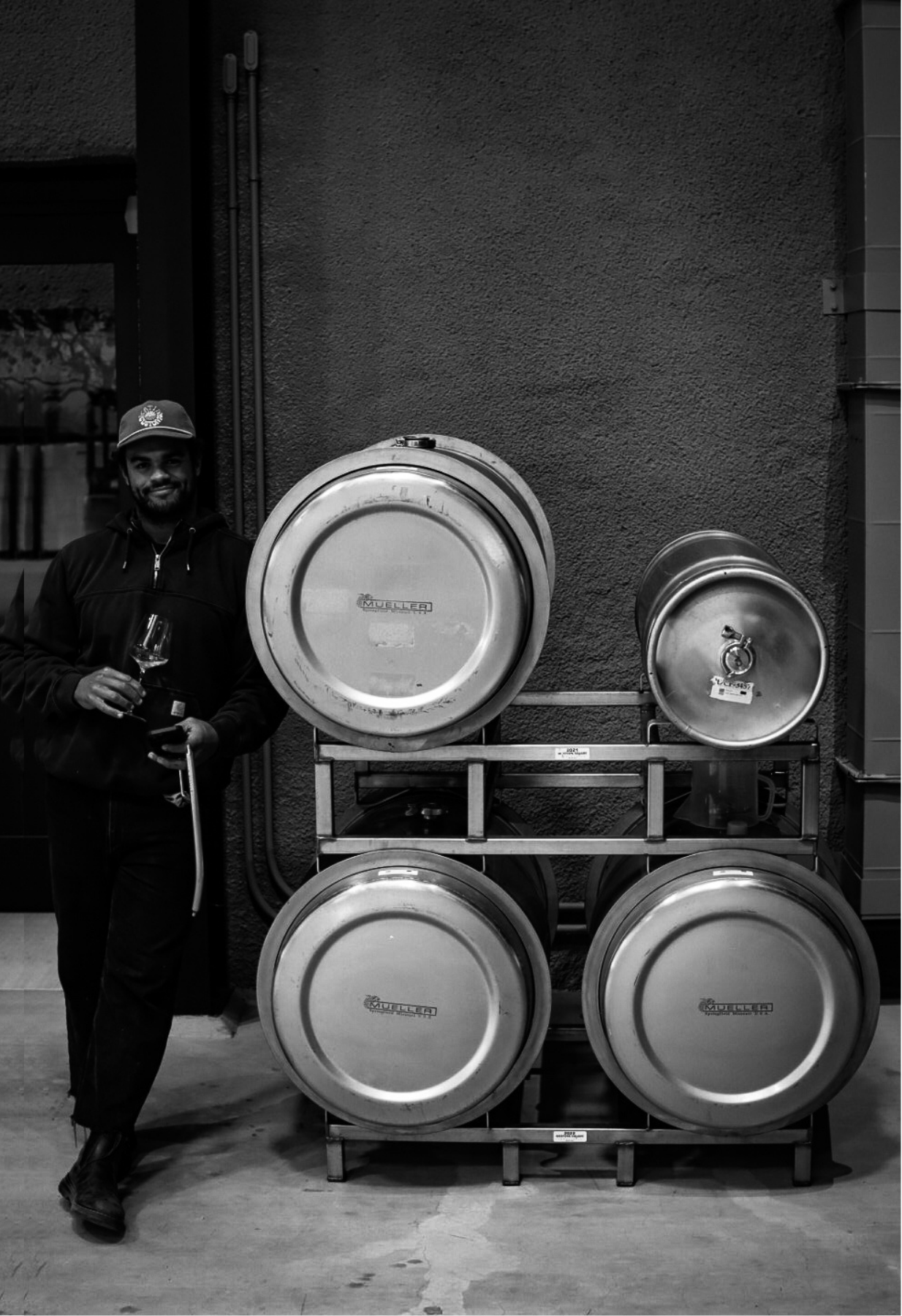 A man stands next to a rack of beer kegs, holding a glass of beer. The image is in black and white.