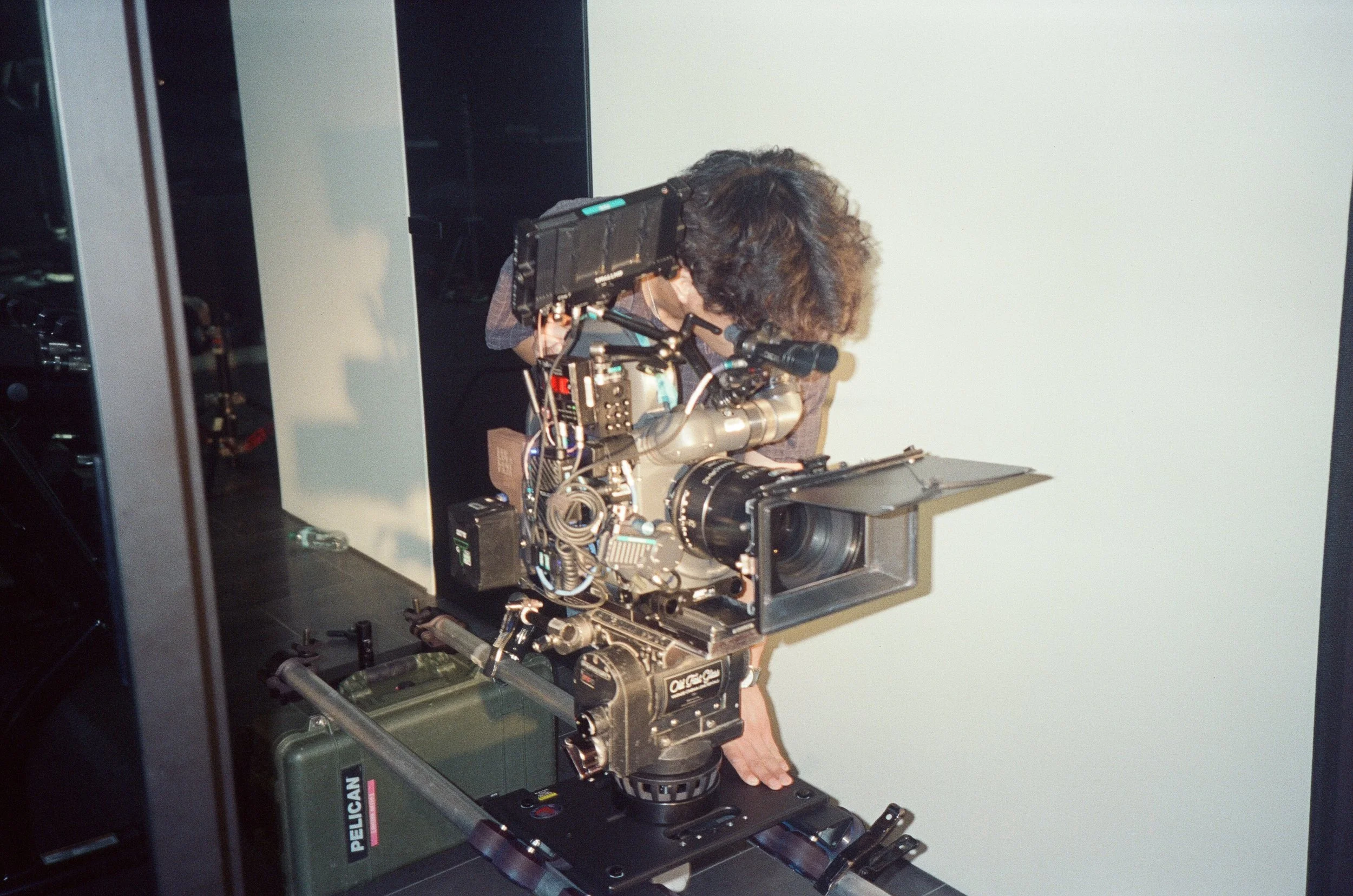 Operator leaning into the ARRIFLEX on a dolly track, matte box forward, beside a glass wall.