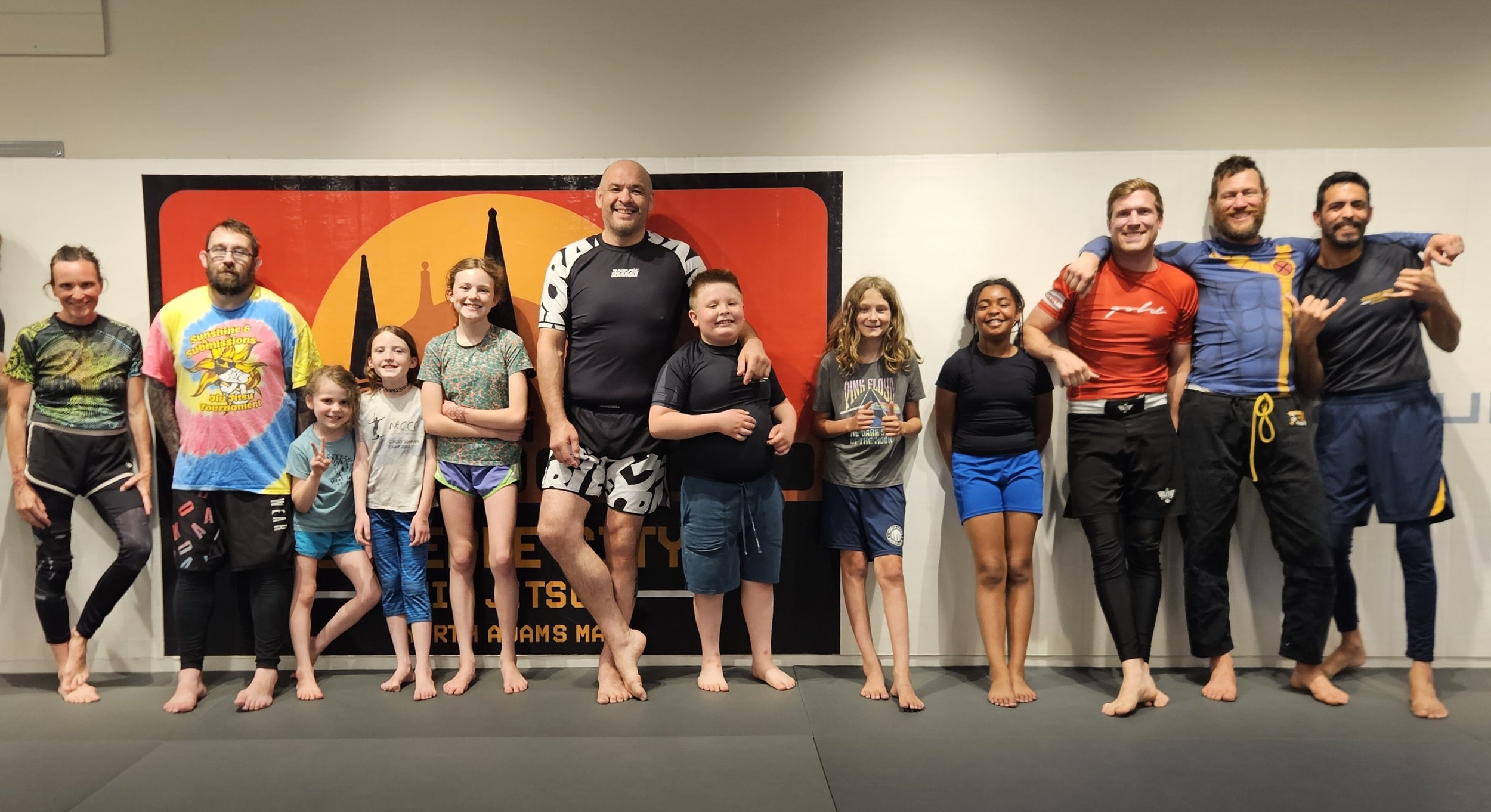 Group of children and adults standing on a martial arts dojo floor, smiling and posing for a photo, with a banner in the background.