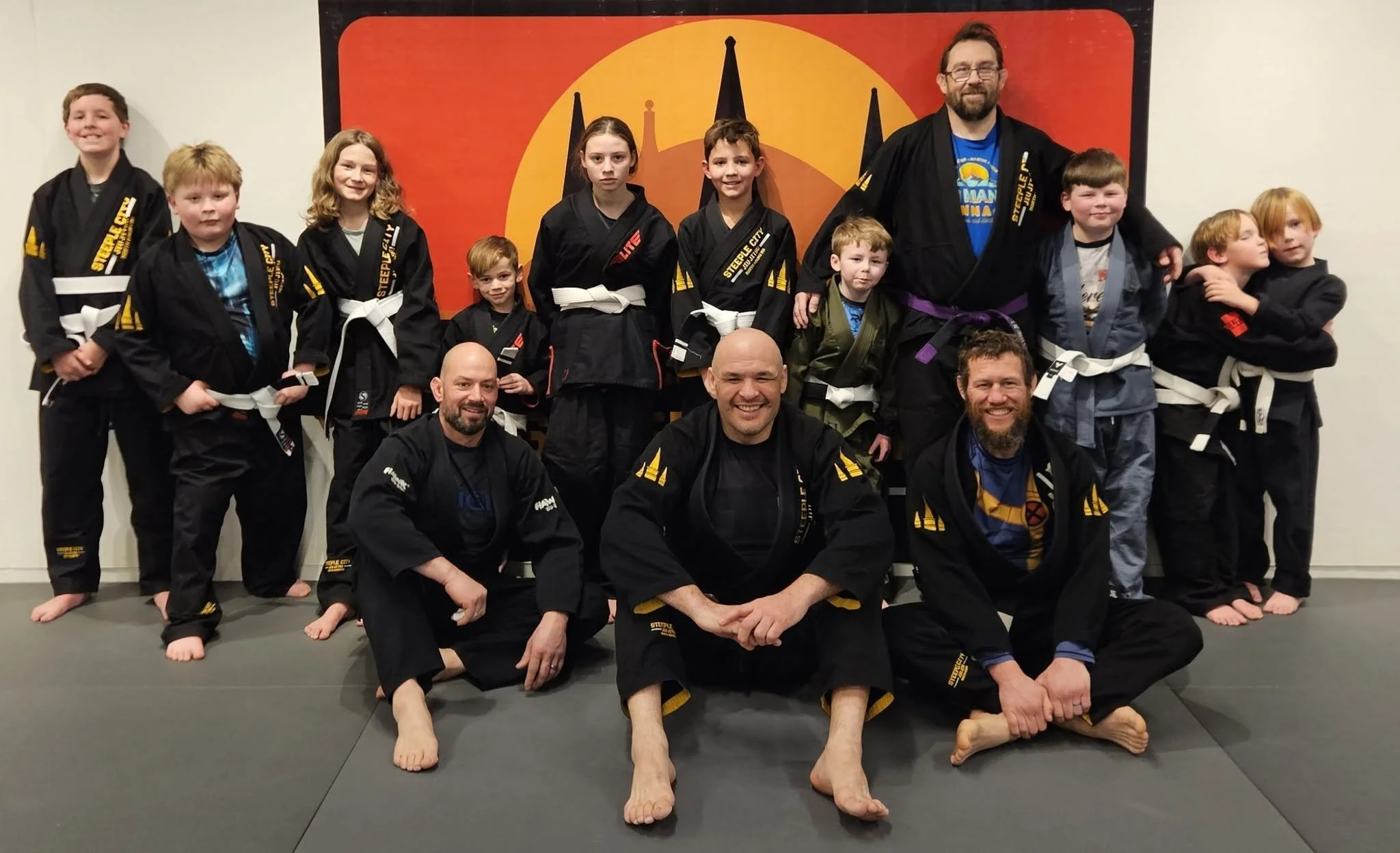 A group of children and three adults in Brazilian Jiu-Jitsu uniforms posing together in a dojo with a red and yellow wall art in the background. Some children are standing, kneeling, or sitting on the mat, and some are hugging or leaning on each other.