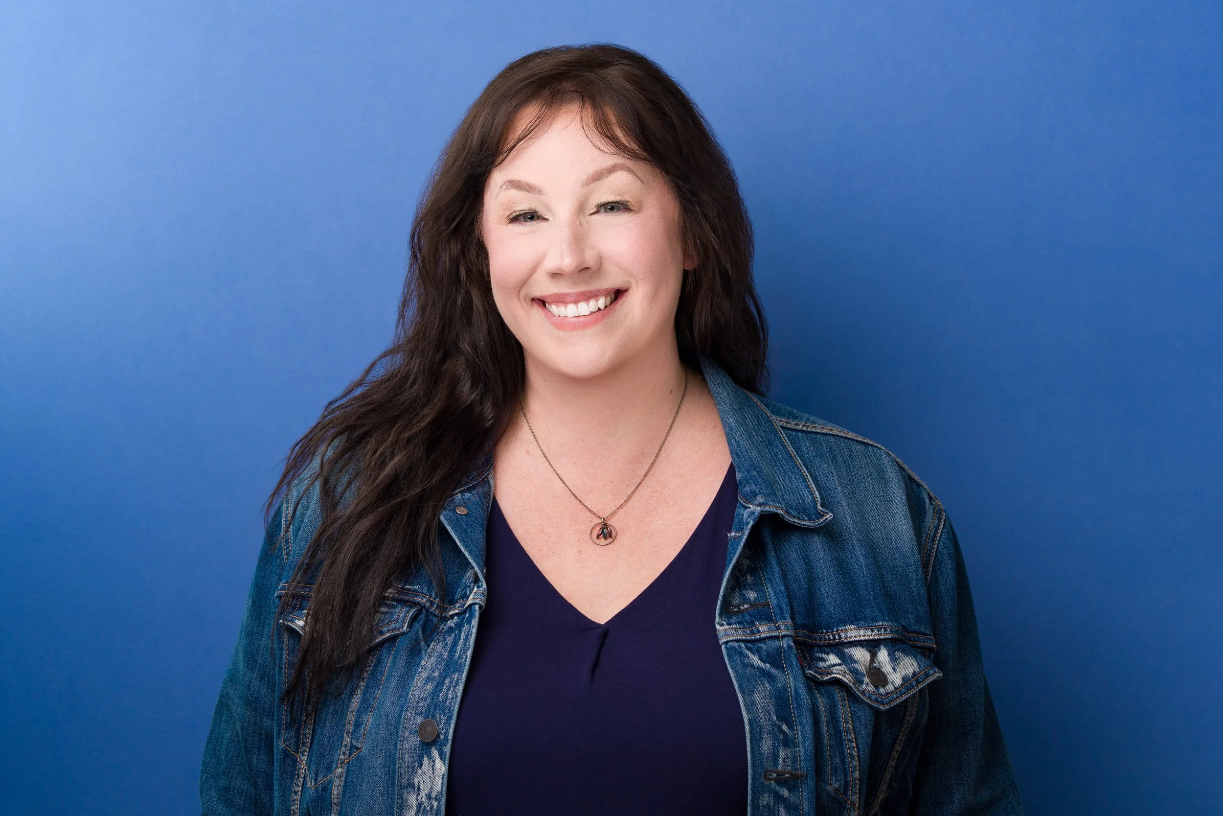 A picture of me smiling with a blue background. I'm wearing a dark blue shirt with a denim jean jacket