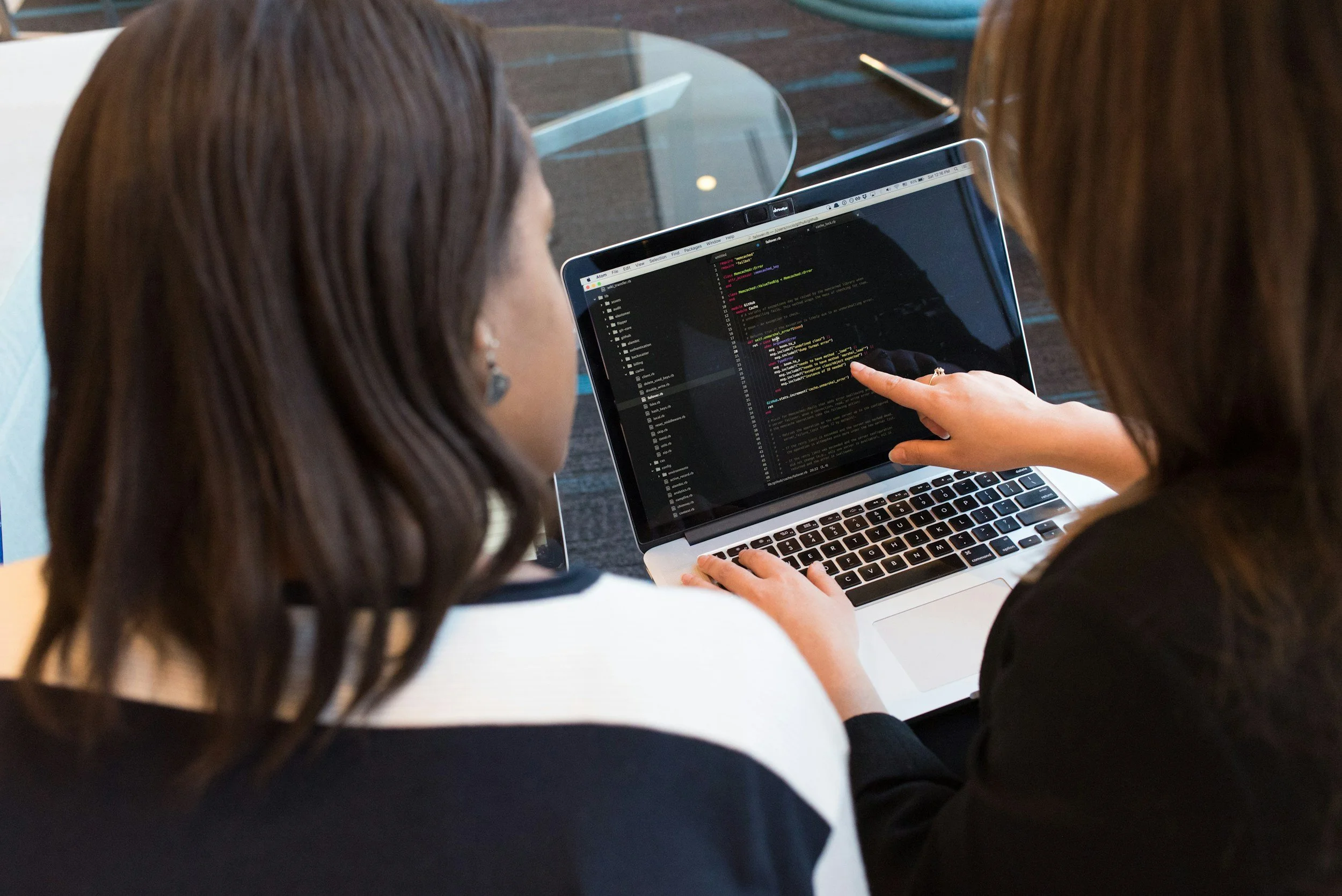 Two women working together on a laptop displaying code. One woman is pointing at the screen, and they are sitting at a table in a modern workspace.