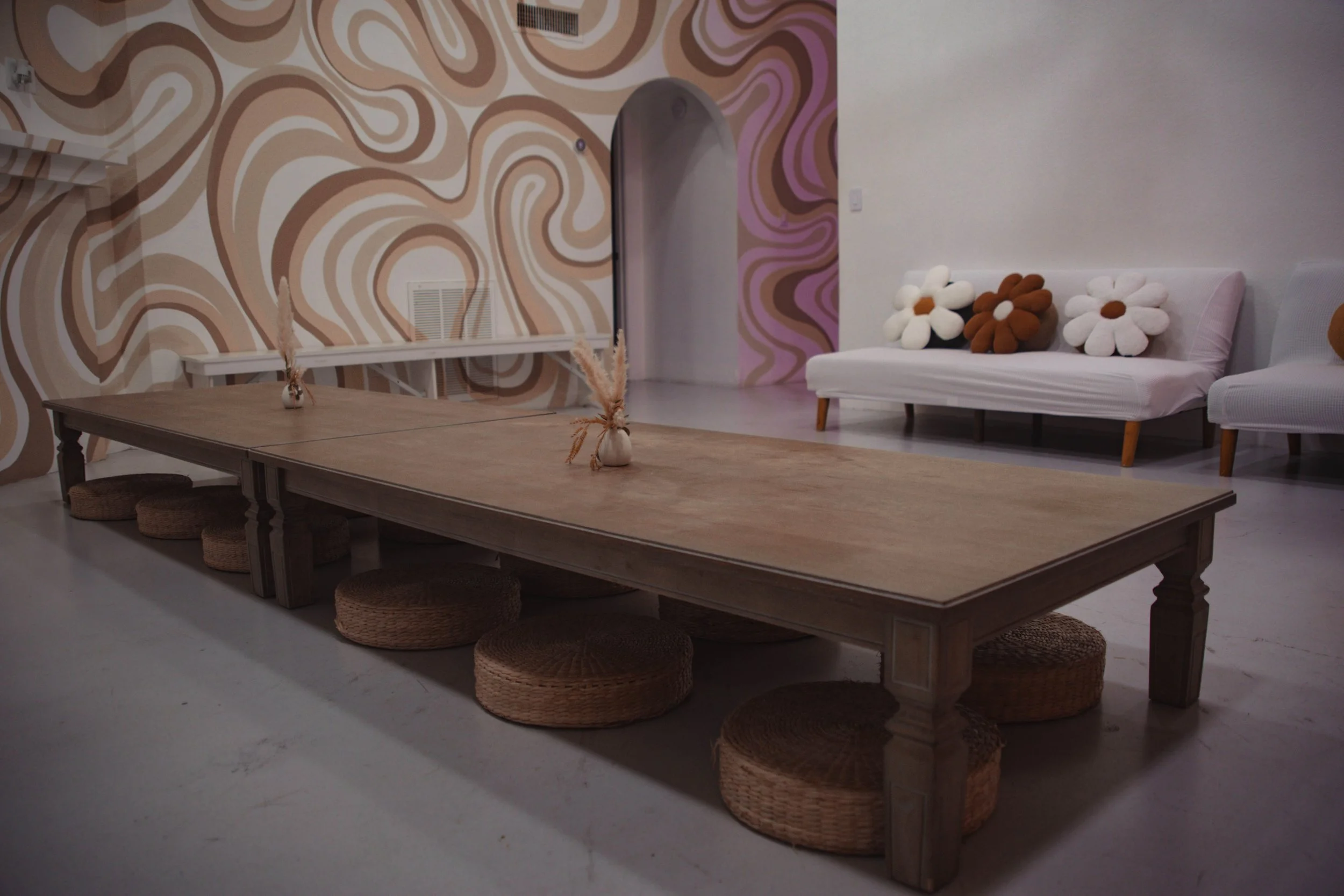 A large wooden table with woven round cushions underneath, two vases with dried plants on top, a patterned wall with swirling designs, and a seating area with a white sofa decorated with flower-shaped pillows in white, brown, and black.