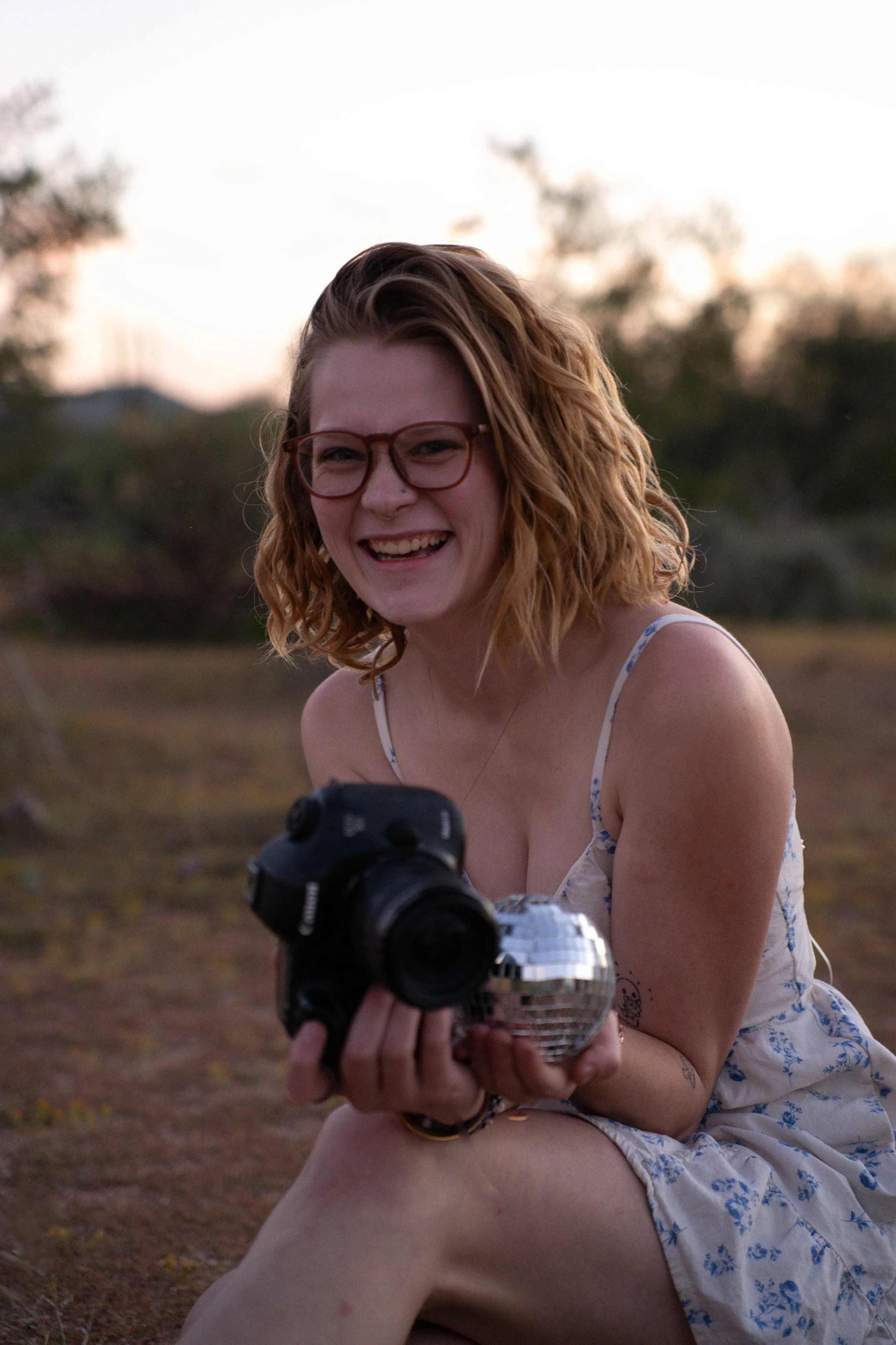 A woman with red hair, glasses, and a floral dress sitting outdoors at sunset, holding a camera and a small disco ball, smiling.