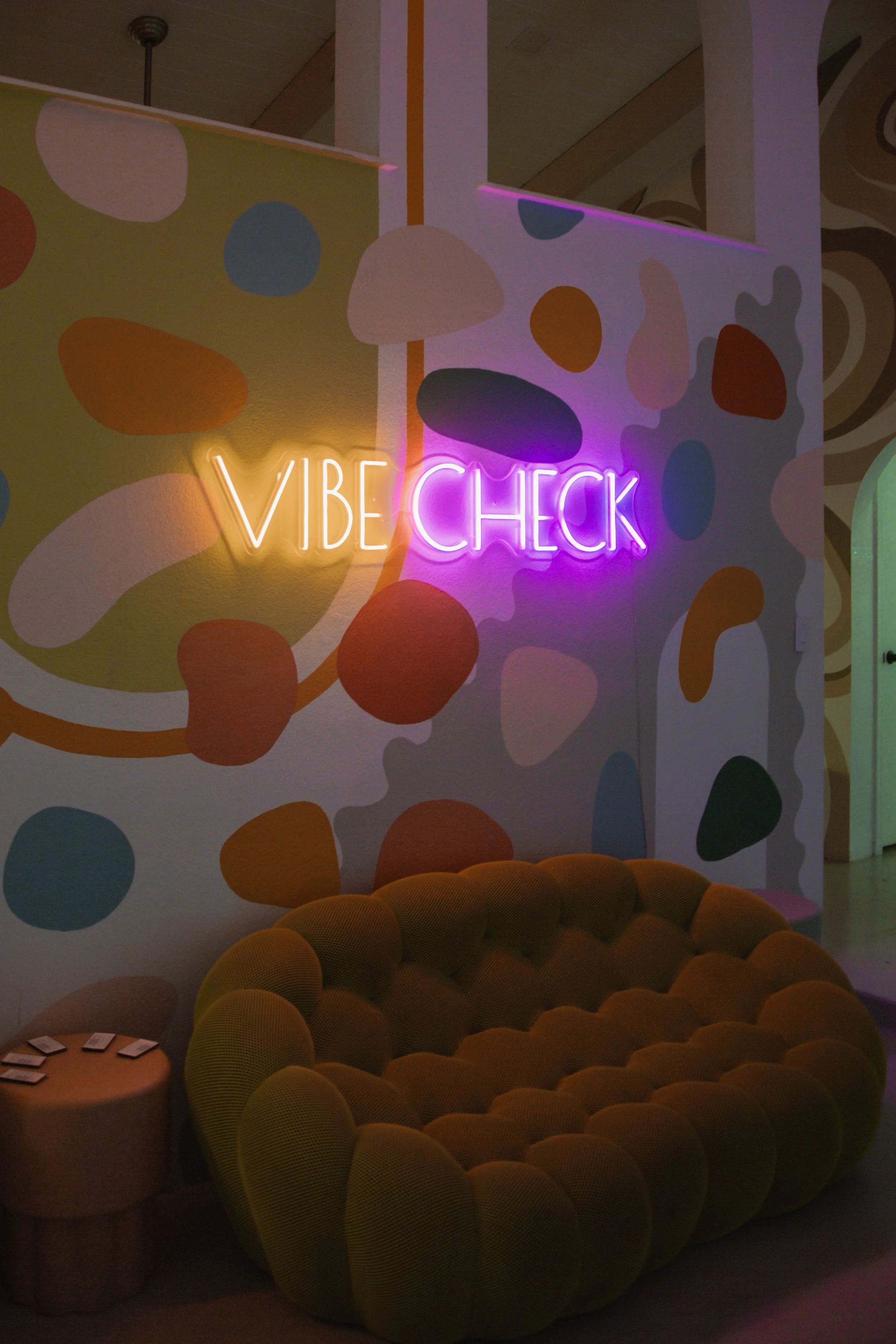 Neon sign reading 'VIBE CHECK' in colorful, modern decor with abstract mural and a plush, circular yellow couch.
