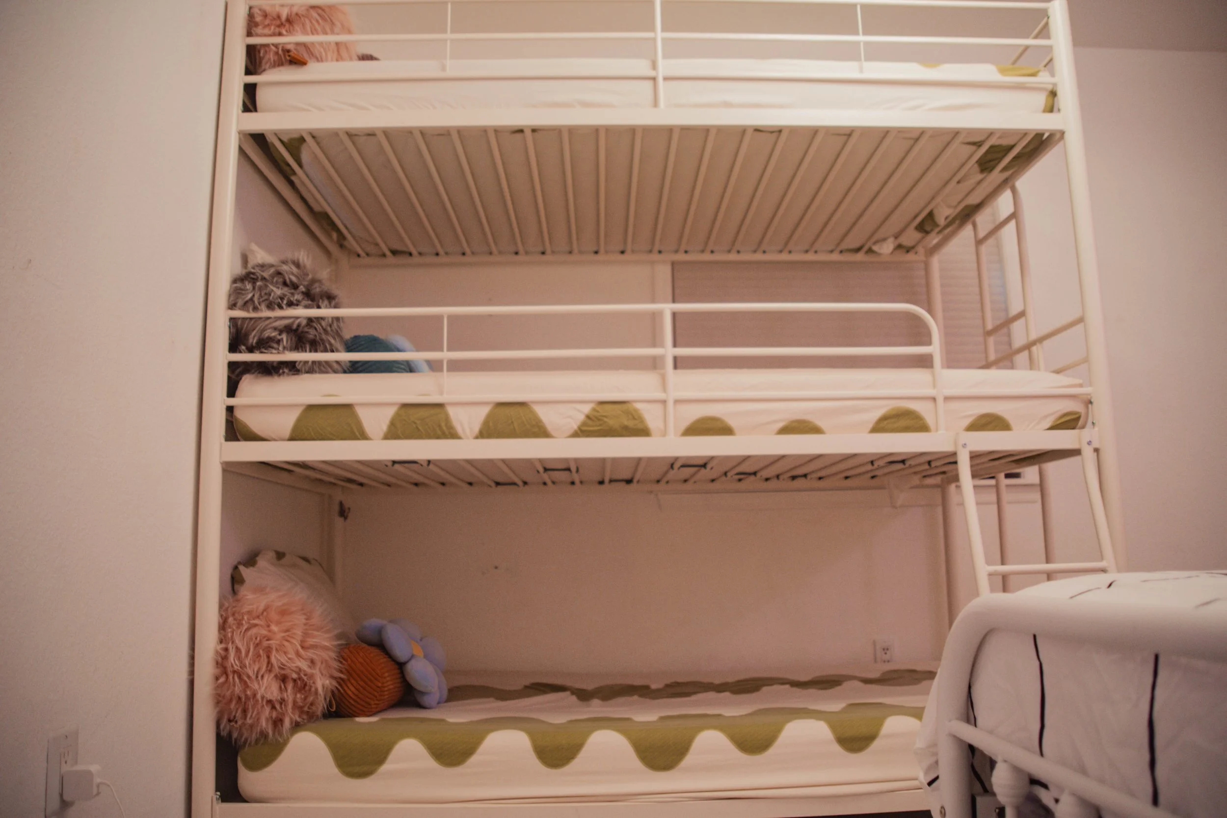 A white metal bunk bed with two levels, each with a mattress and bedding. The bottom bunk has a pink fluffy pillow, a plush flower, and a brown striped plush pillow. The top bunk has a pillow and blanket, with a pink fluffy pillow visible in the corn
