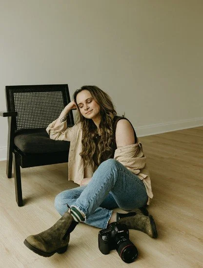 A young woman with long wavy hair in a beige top and jeans, sitting on the floor next to a camera, leaning against a black chair in a room with light-colored walls and wooden flooring.