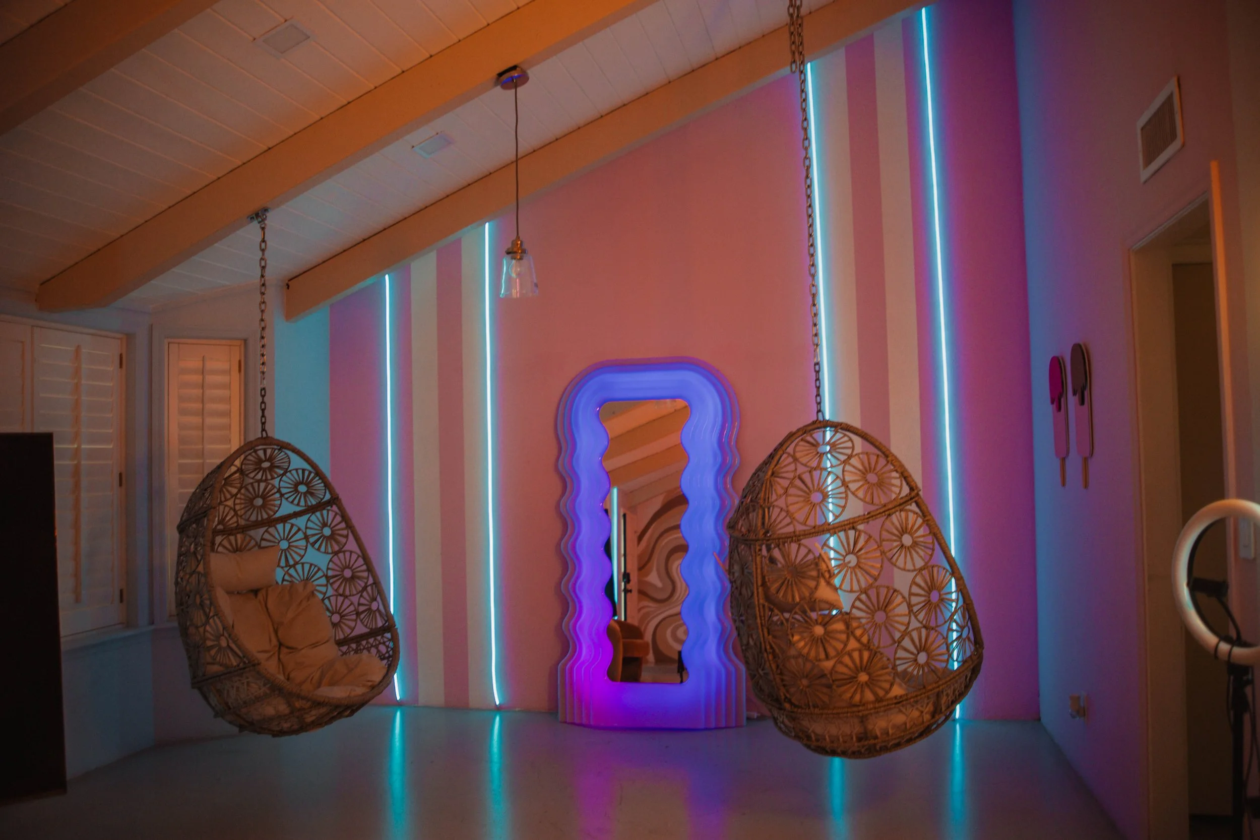 A room with hanging wicker egg-shaped chairs, neon blue and pink vertical lights on a striped pink and white wall, a decorative mirror, and wall art.