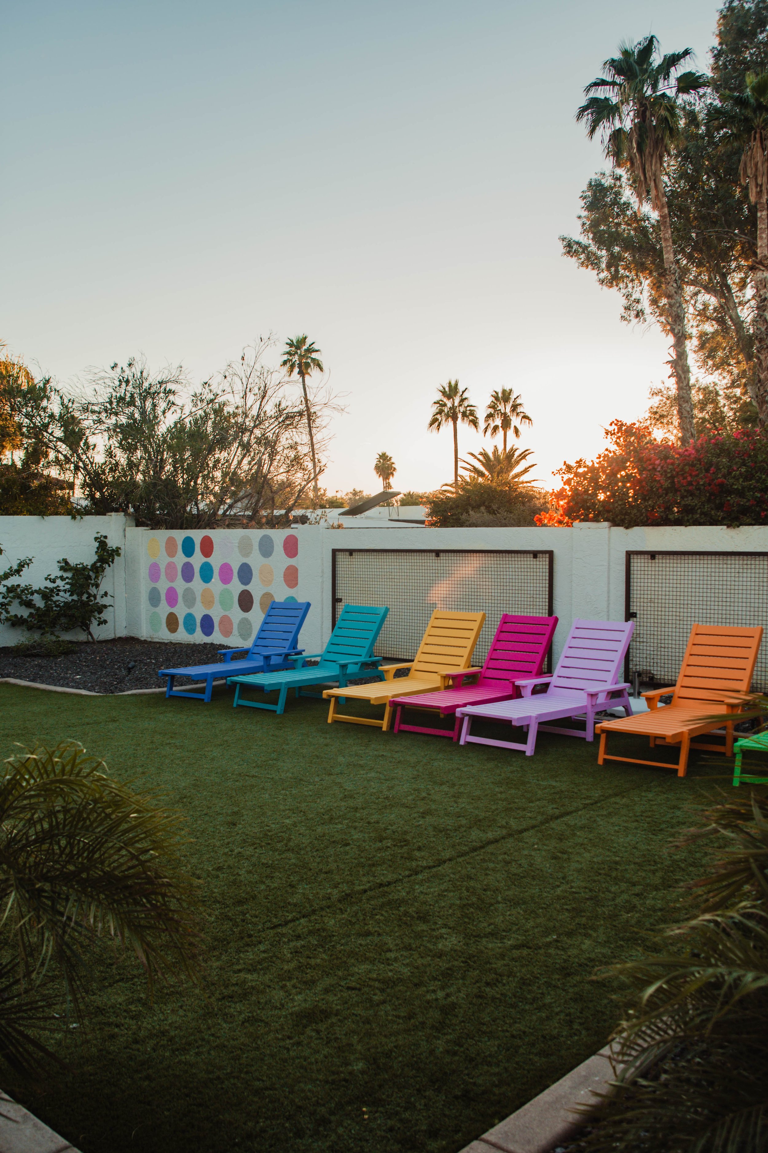 Colorful lounge chairs in blue, teal, yellow, pink, purple, and orange, arranged in a row on a grassy yard with a white fence and decorative wall with polka dots, surrounded by palm trees at sunset.