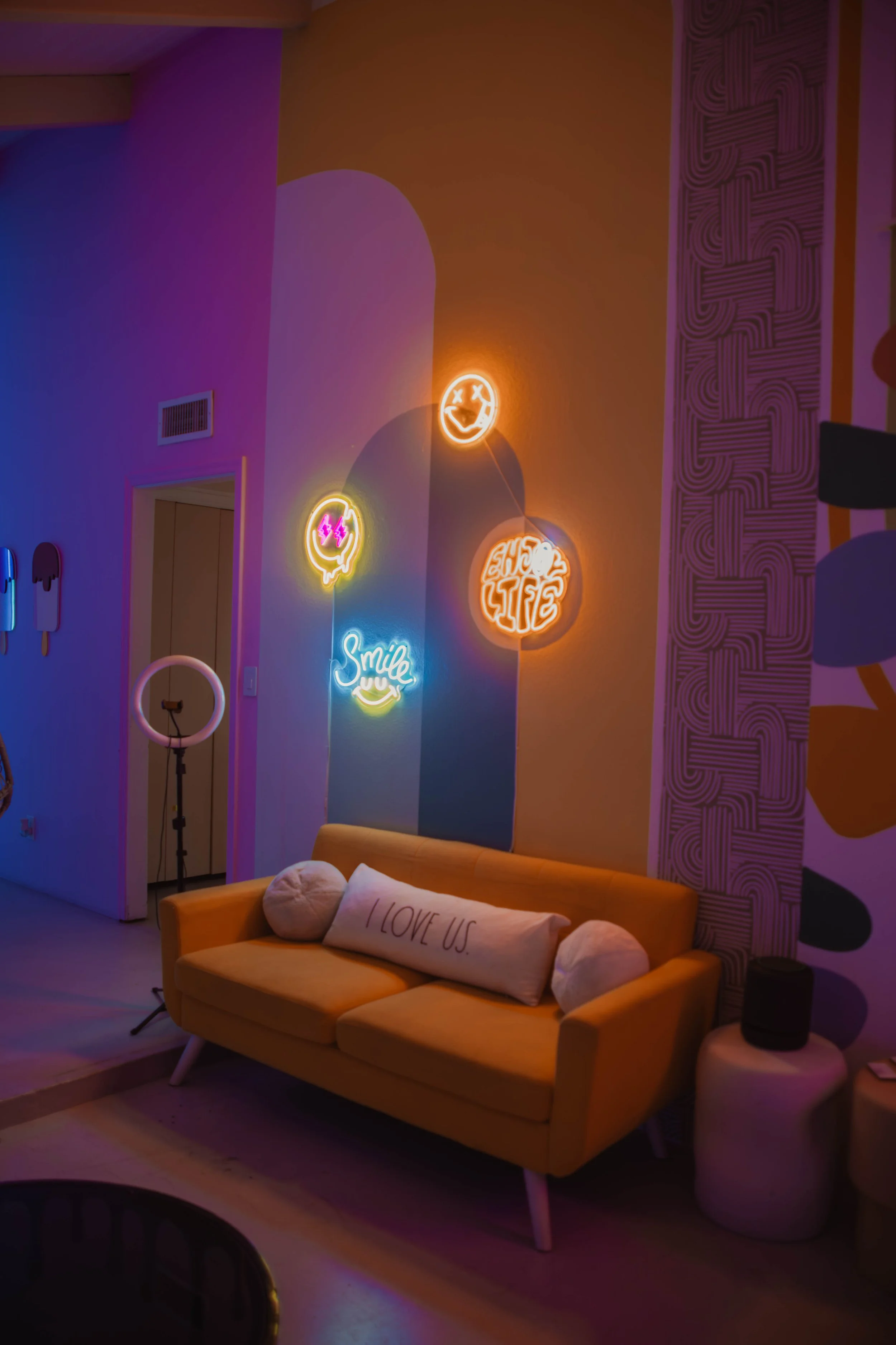Living room with colorful neon signs on the wall, a yellow sofa with a pillow that says "I LOVE US," and a ring light. Neon signs include smiling face, speech bubble with lightning, "SMILE," and "CHILL LIFE." The room has vibrant purple, orange, and 