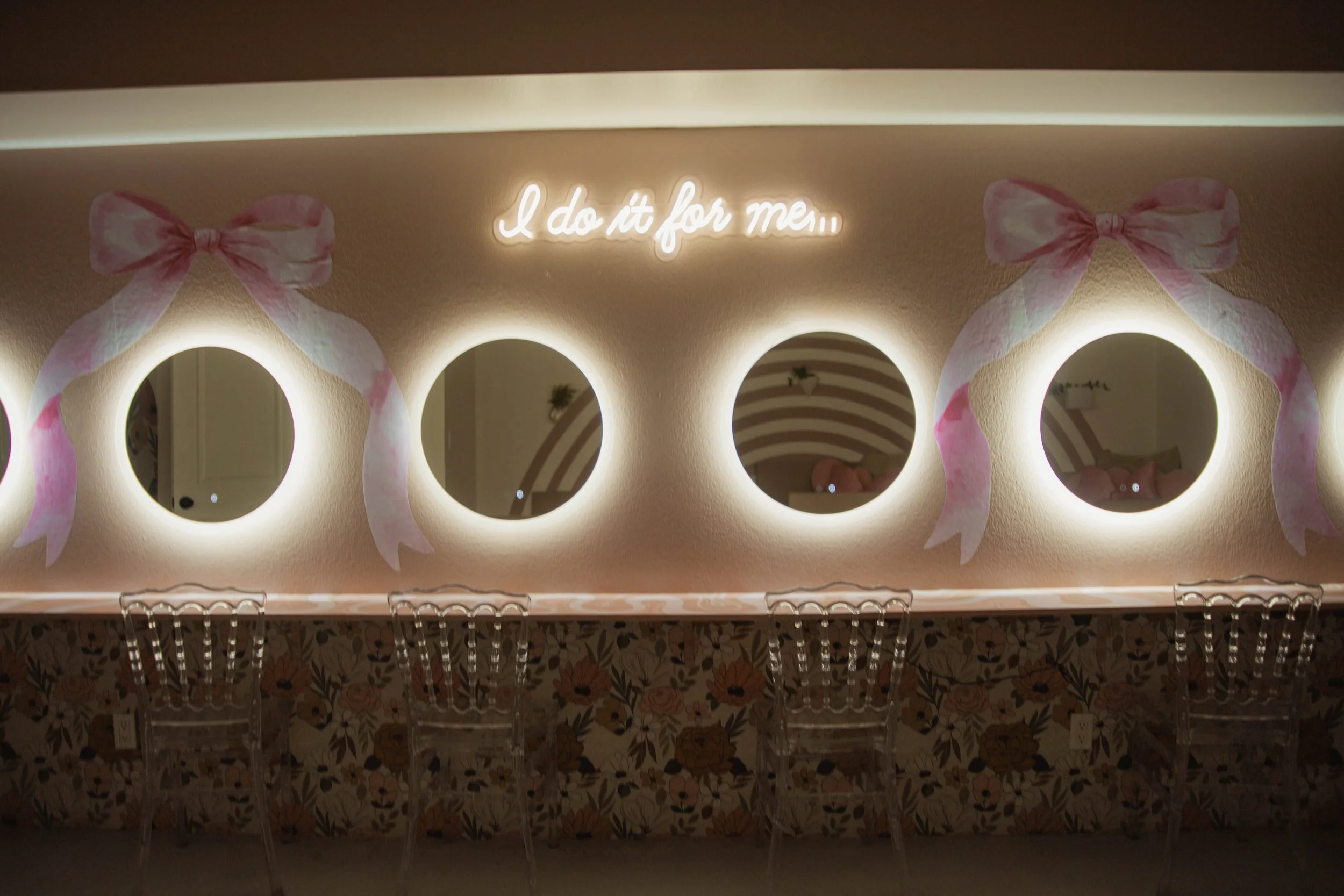 A wall decor with four round mirrors, pink ribbons, and a neon sign that says "I do it for me". There are transparent chairs in front of a floral tablecloth.