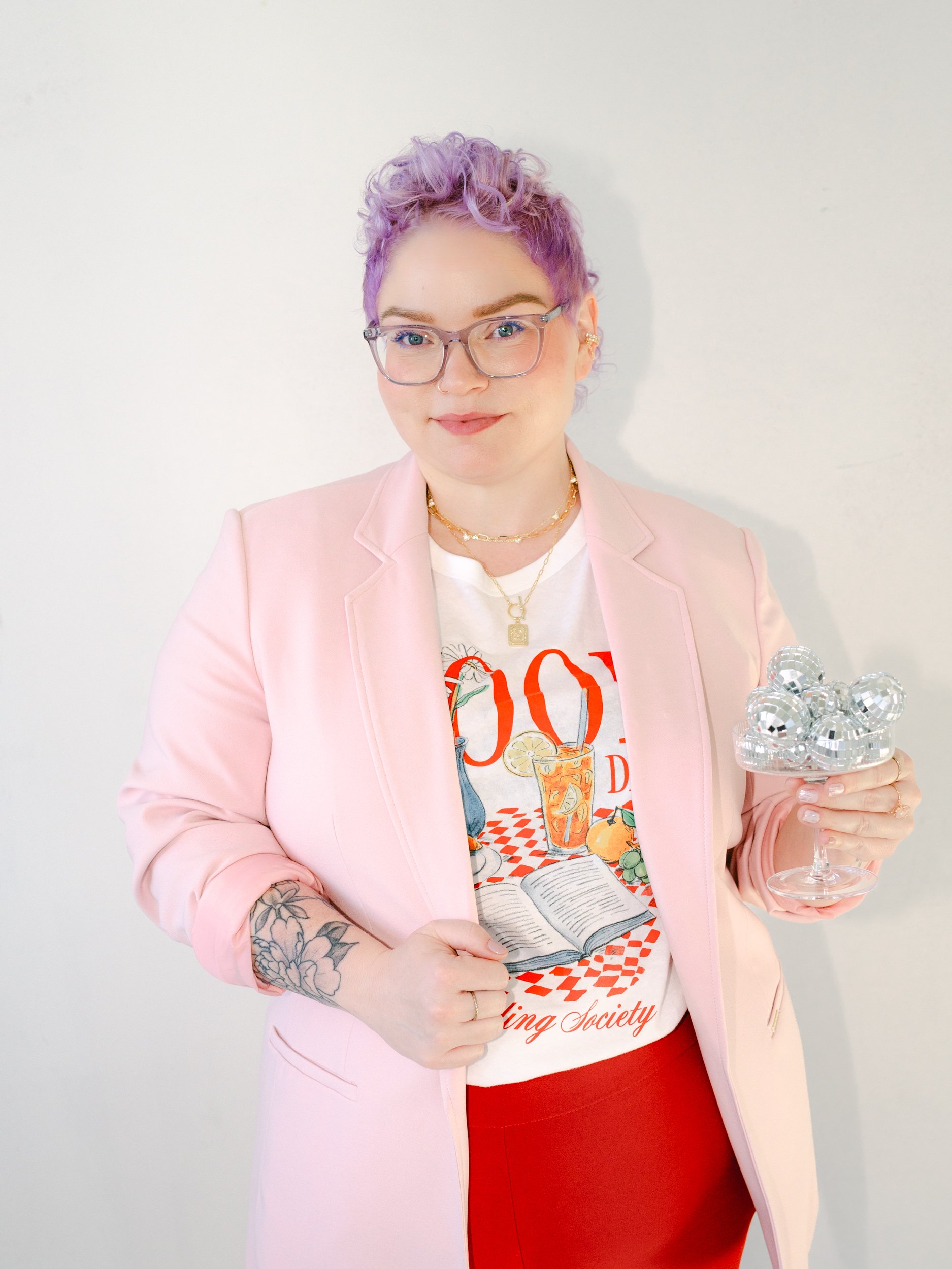 Woman with purple curly hair and glasses, wearing a pink blazer over a colorful graphic T-shirt, holding a glass bowl filled with small silver disco balls.