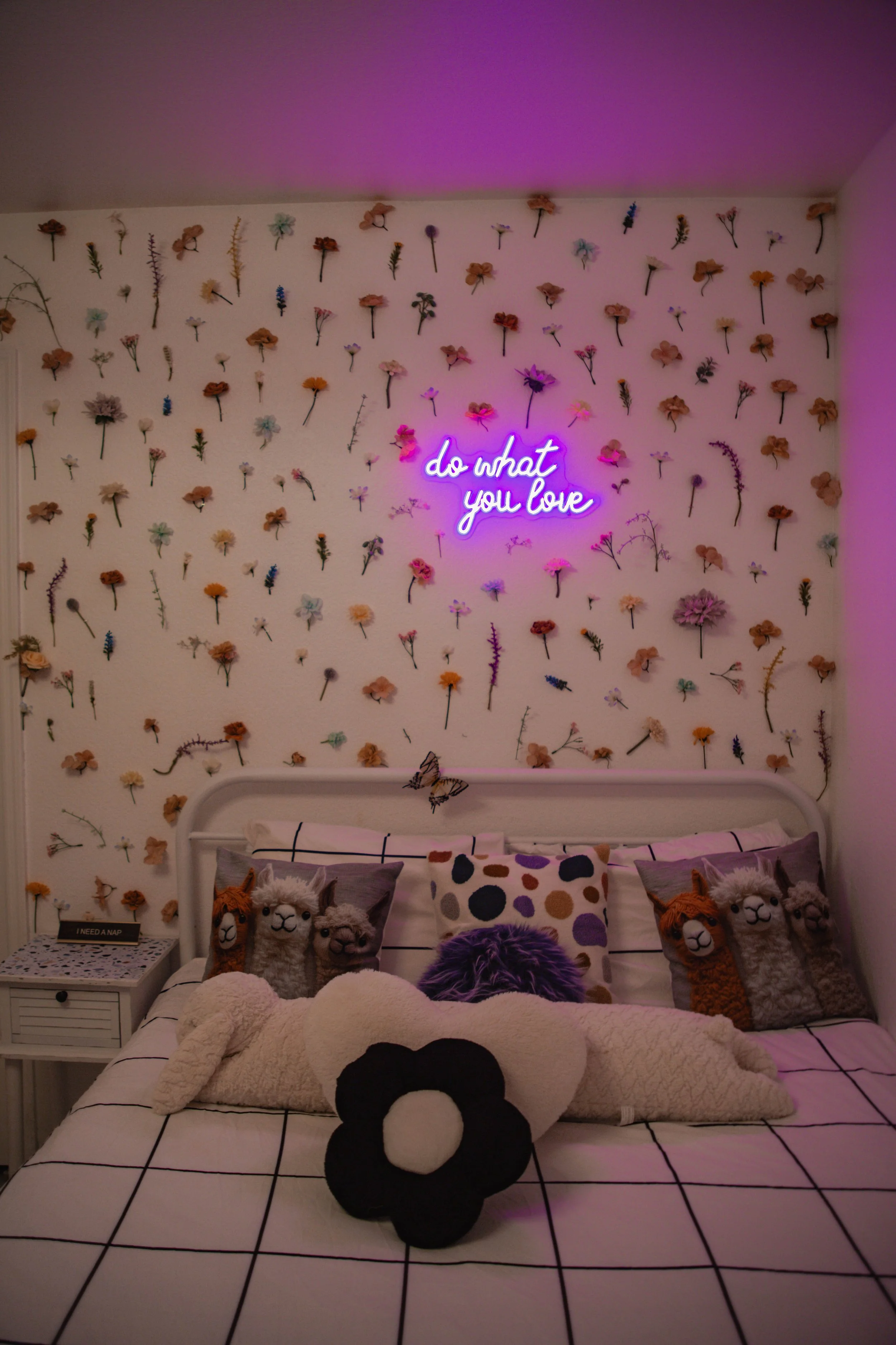 A bedroom wall decorated with dried flowers, a neon sign saying 'do what you love', and a bed with llama plush toys, pillows, and a large flower-shaped plush.