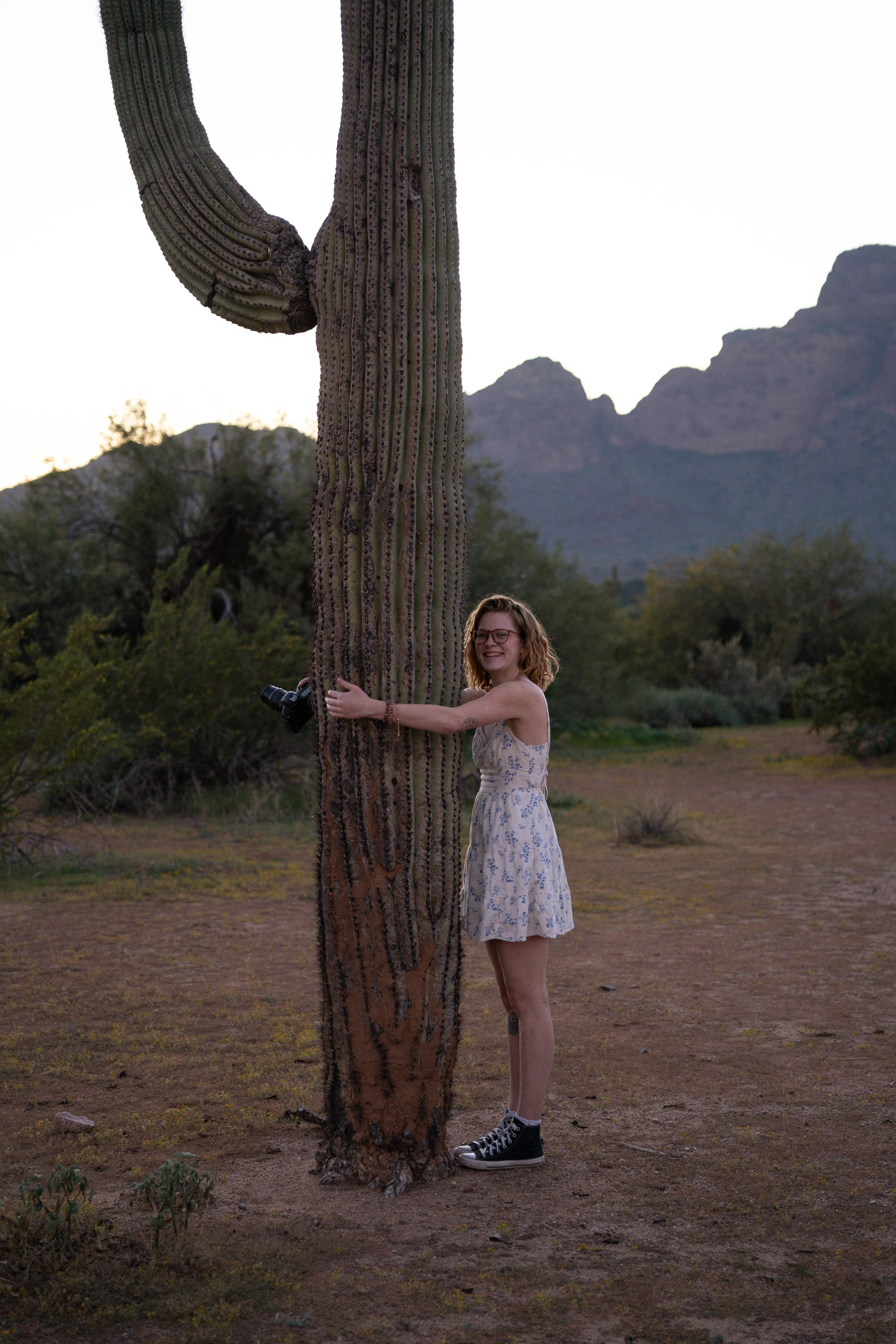 A young woman with glasses, wearing a white dress with blue floral pattern and black sneakers, hugging a tall, large saguaro cactus in a desert landscape during sunset with mountains in the background.