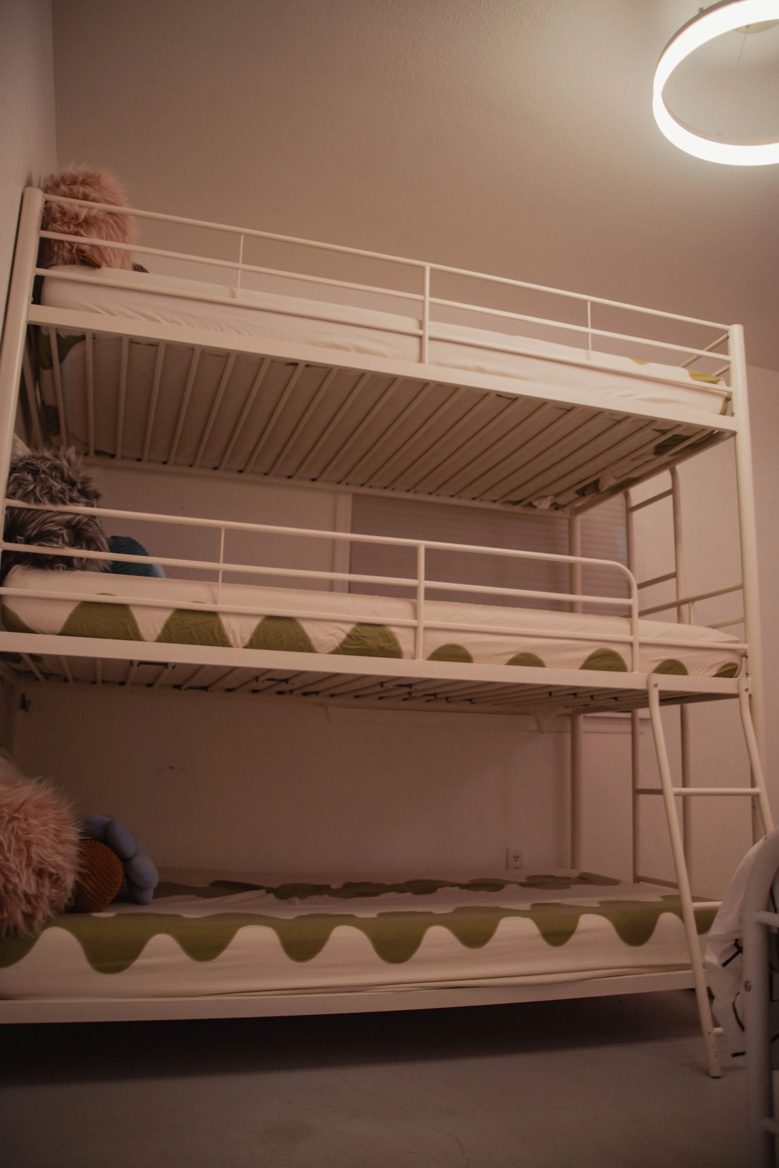 A white metal bunk bed with three levels in a room with a beige wall and ceiling light. The mattresses have green and white triangular patterned sheets, and there are stuffed animals on the bottom and top beds.