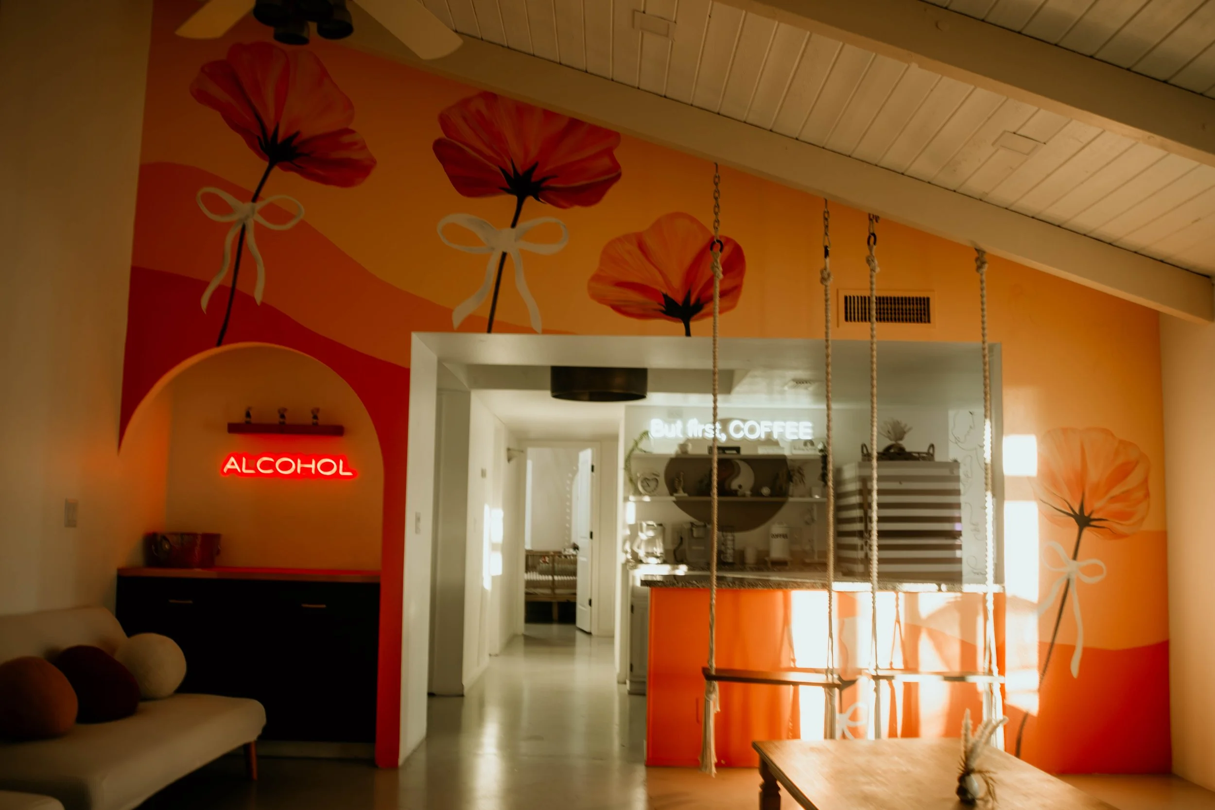 Interior view of a cozy cafe with a vibrant orange wall decorated with large floral murals, a neon sign reading 'ALCOHOL' and a partially visible soft white couch with round cushions.