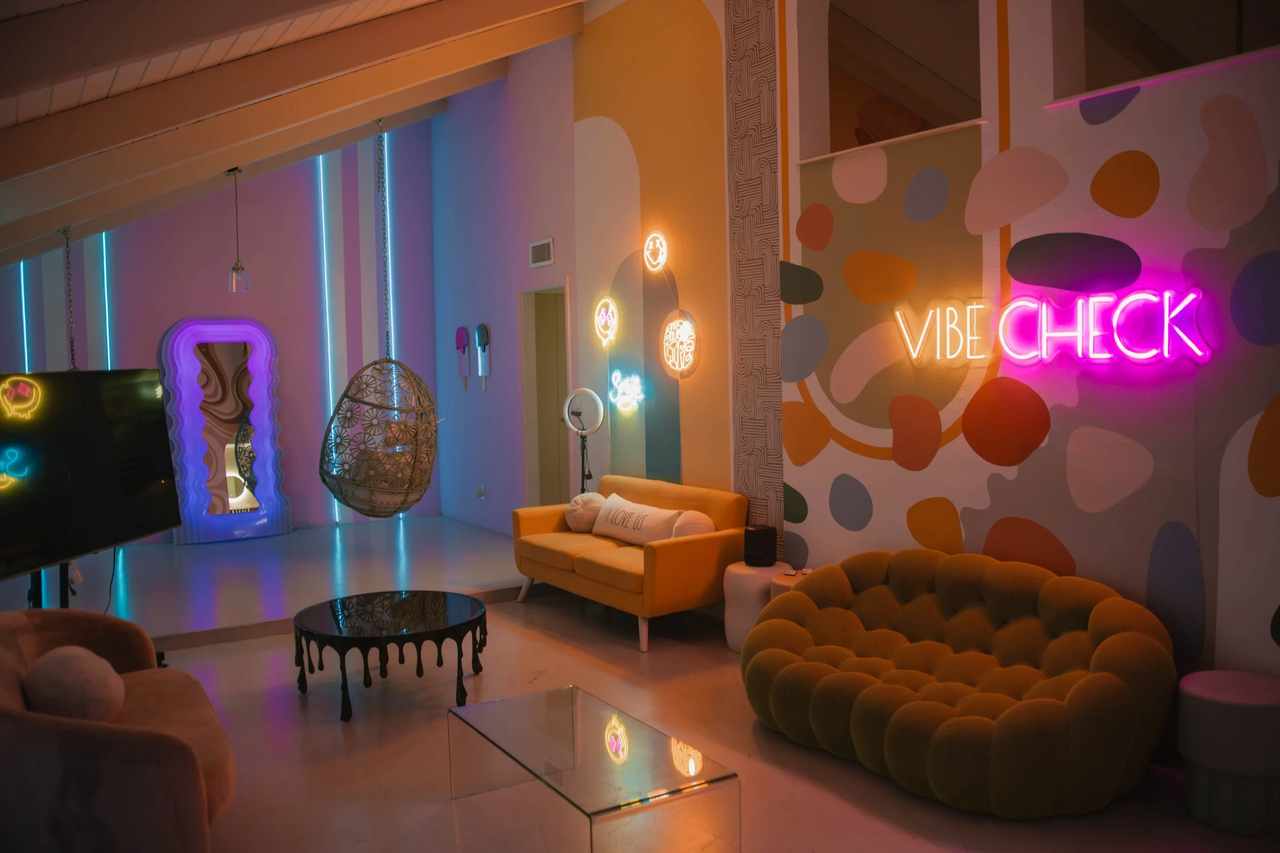 Colorful, neon-lit lounge with various seating options, including a yellow couch, a green velvet circular sofa, and a peach-colored armchair. There is a black, drippy coffee table, a hanging wicker chair, and vibrant wall art with neon signs that say