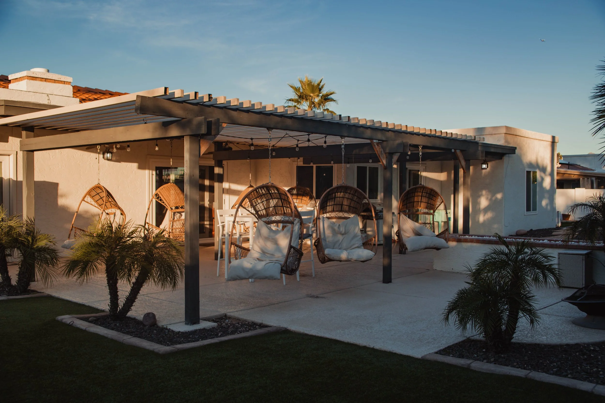 A cozy backyard patio with hanging wicker egg chairs, a white table and chairs, and small potted palm trees. The patio has a pergola with string lights, and the house is white with windows. The sky is clear and blue, suggesting evening or late aftern