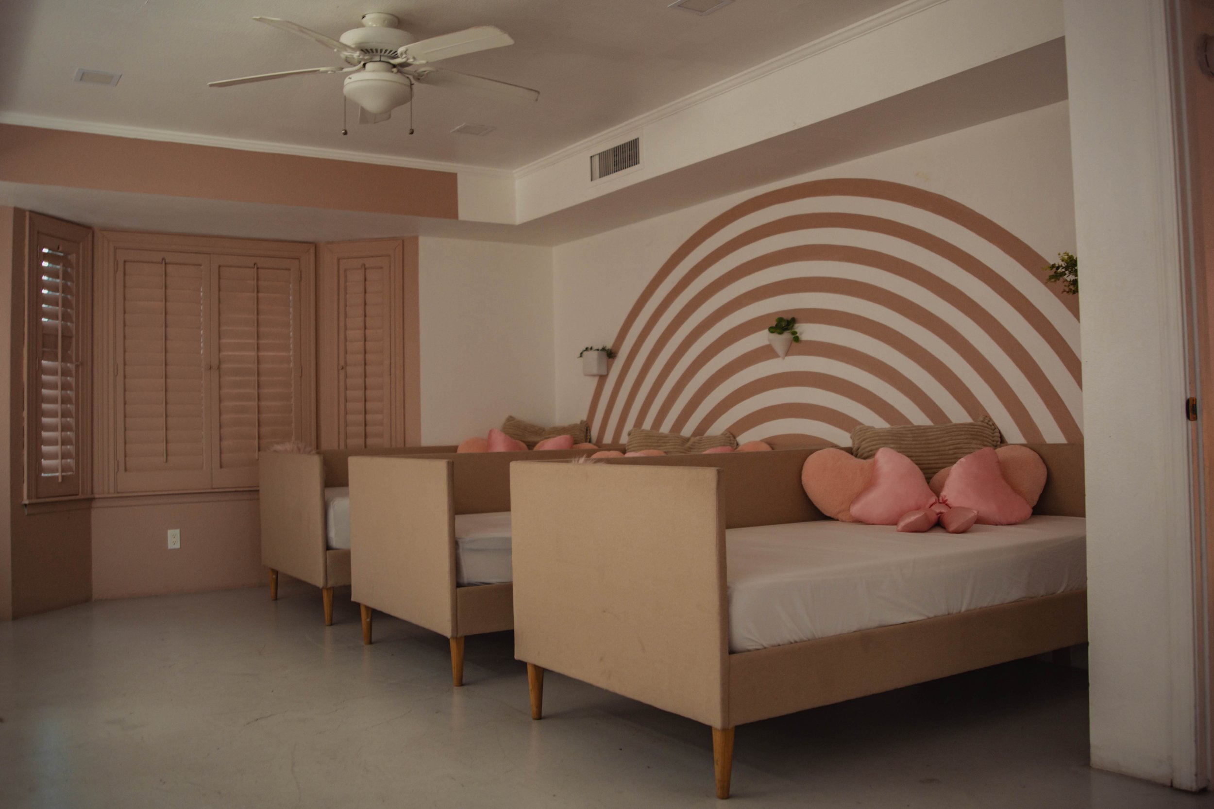 A cozy living room with beige sofas, pink heart-shaped and pillow-shaped cushions, a white ceiling fan, and a wall with pink and white rainbow stripes and small wall planters.