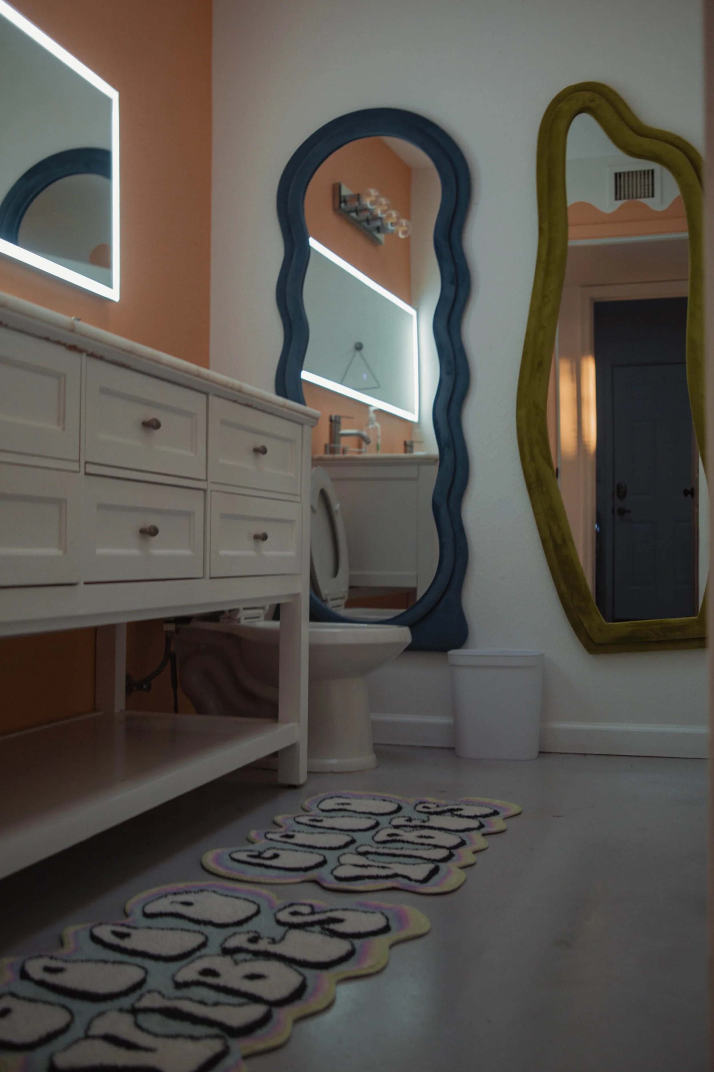A bathroom with a white vanity, two mirrors with colorful frames, a closed toilet, and a colorful mat on the floor that reads 'BUBBLES'.