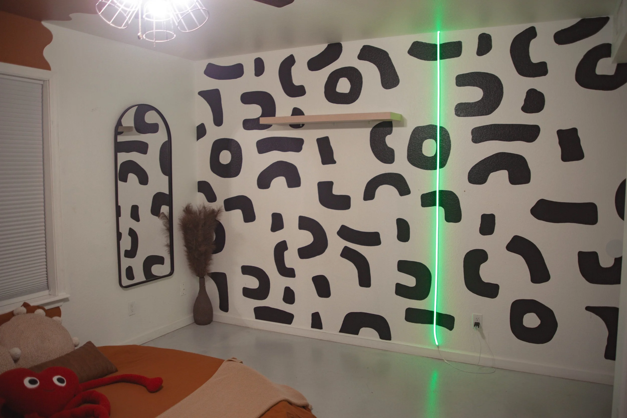 Bedroom with a black and white abstract patterned accent wall, a black mirror frame, a floor-length mirror, a tall vase with furry pampas grass, a bed with red and brown plush toys, and a green LED light strip on the wall.