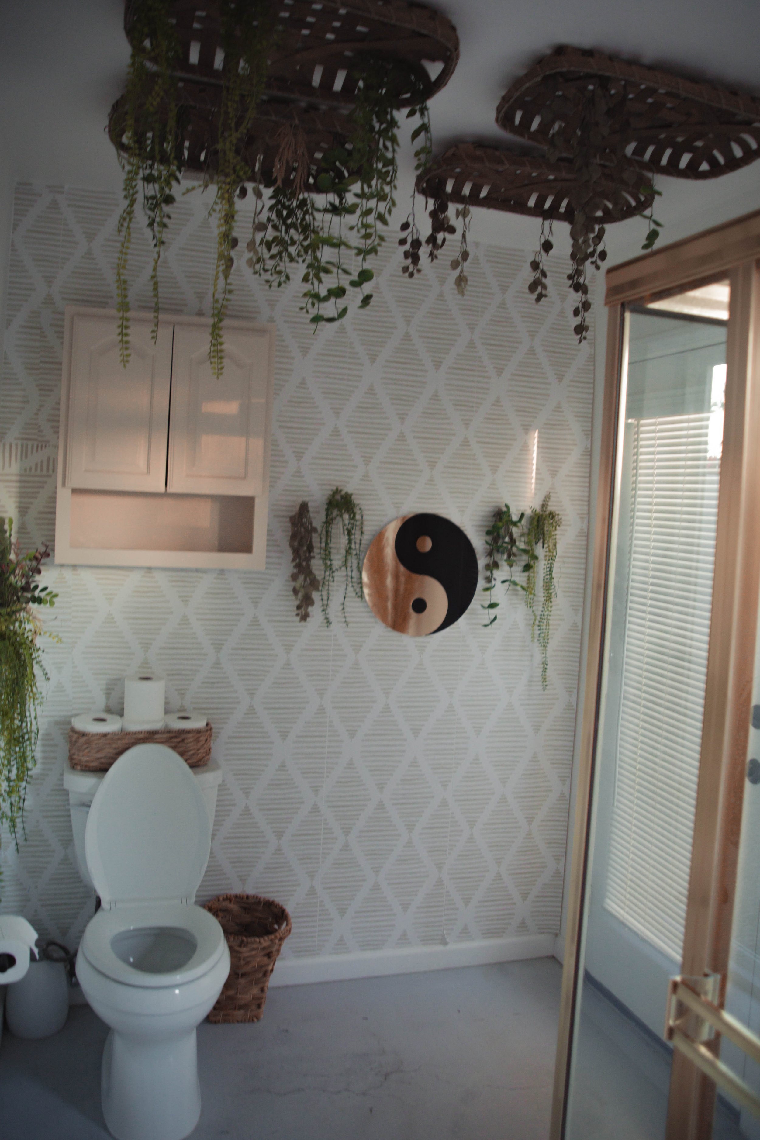 A bathroom with textured wallpaper, hanging plants, a yin-yang wall decoration, a toilet with a basket on top, and a sliding door.