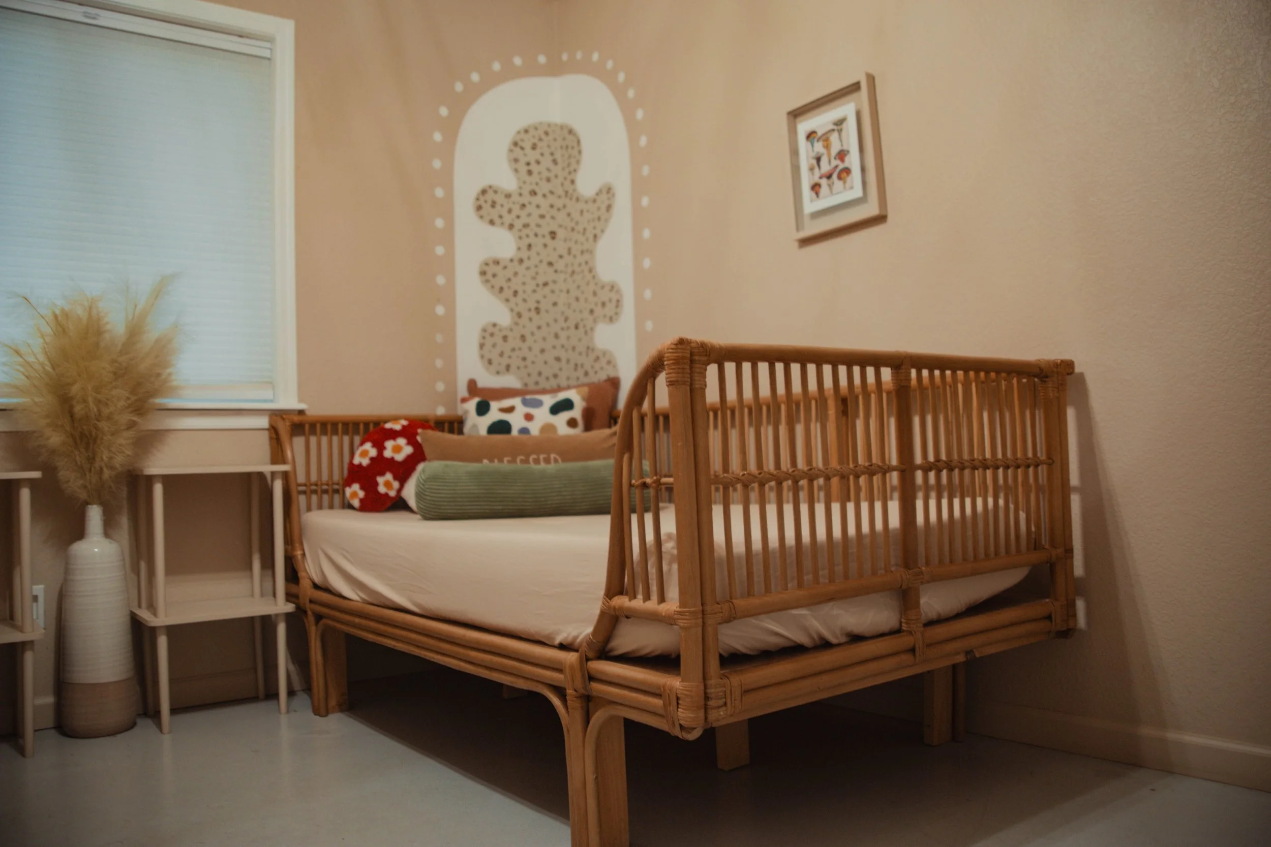 A bedroom with a rattan bed, decorative pillows, wall art, a window with a blind, and a large vase with dried pampas grass.