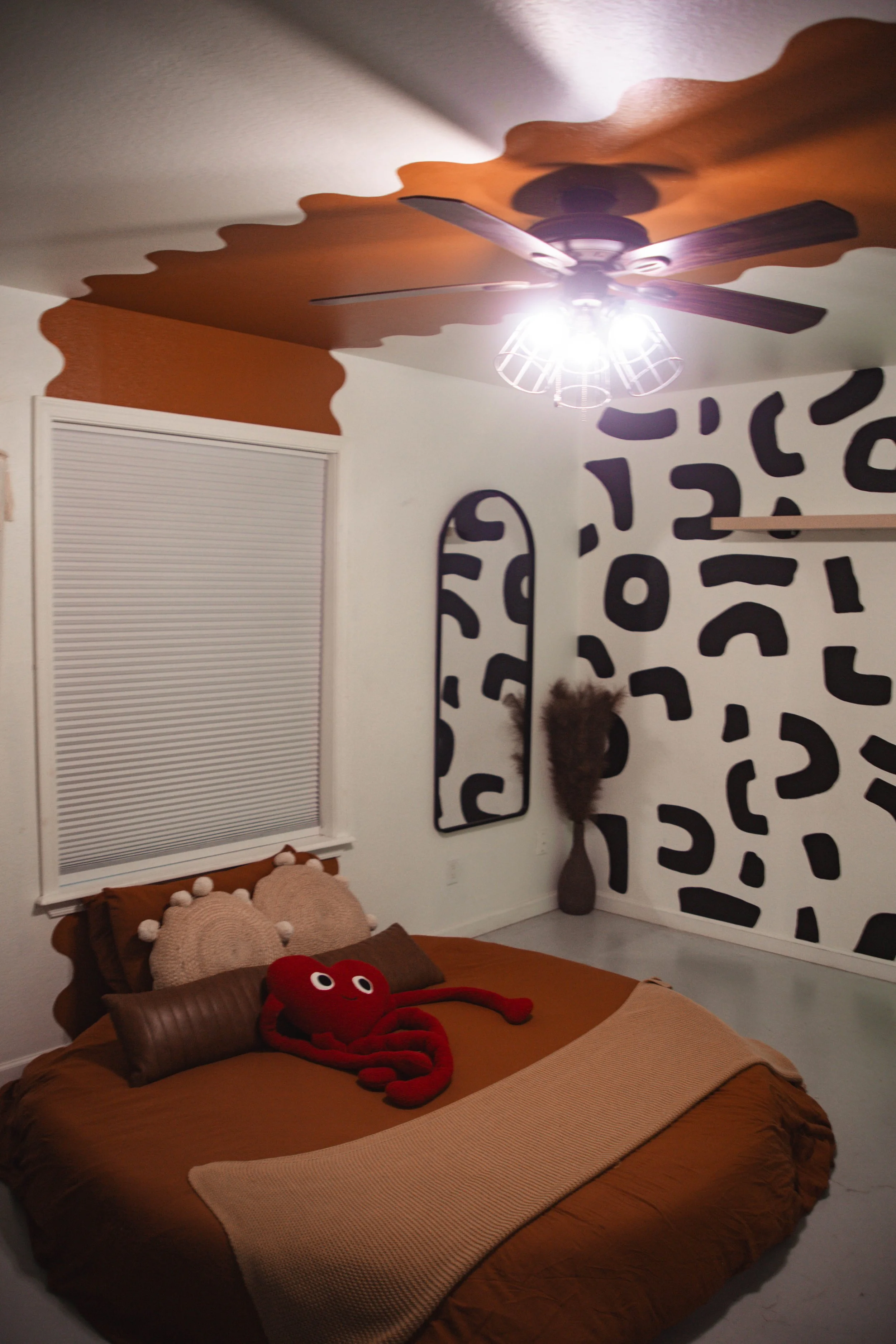 A bedroom with a low bed covered in brown bedding and pillows, one pillow resembling a bear, and a plush red octopus toy on the bed. The wall has abstract black patterns, and a mirror and tall vase with dried plants are in the corner. The ceiling fan