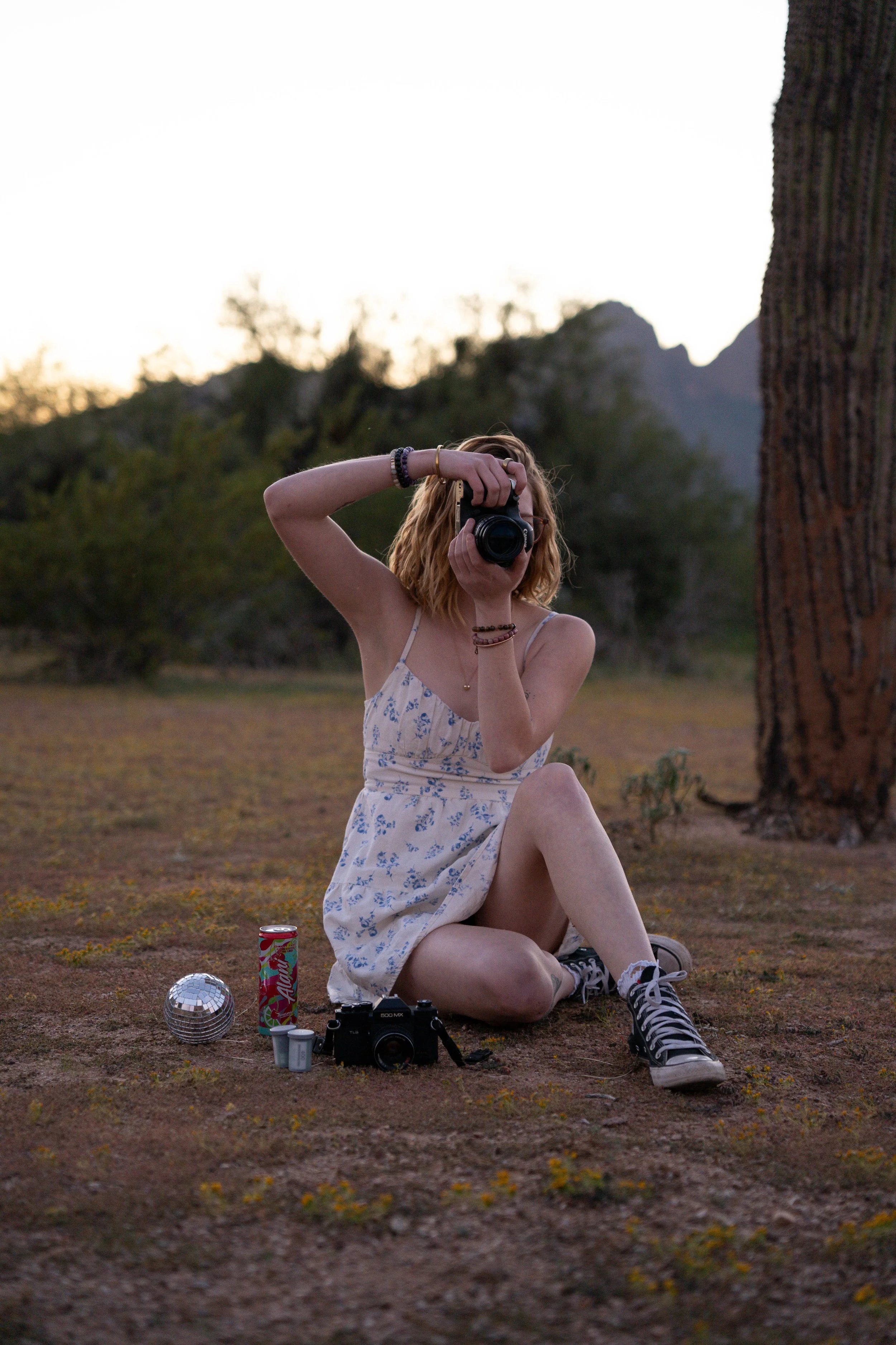Young woman in a summer dress sitting on the ground outdoors at sunset, holding a camera to her face, taking a photo. There is a camera on the ground beside her along with a soda can, a disco ball, and a small container. Background features distant trees and mountains.
