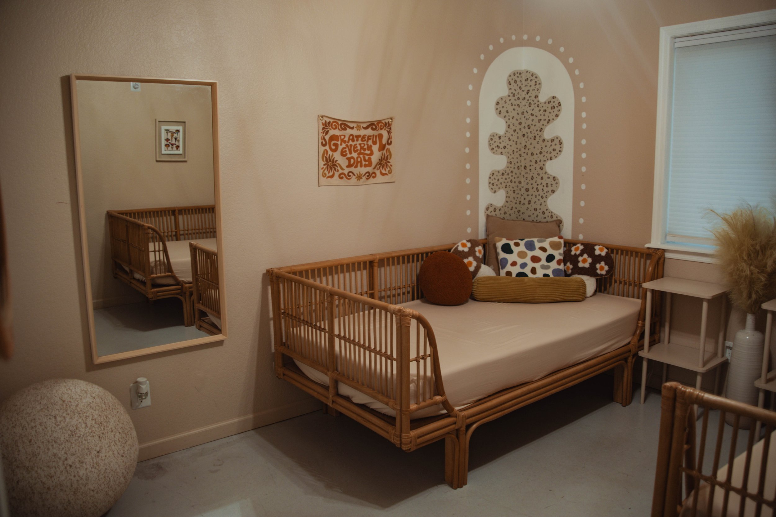 A cozy bedroom with a rattan daybed adorned with colorful pillows, a large mirror on the wall reflecting the bed, a wall decoration with the phrase 'Grateful Every Day,' a small side table, and a vase with dried pampas grass next to a window with bli
