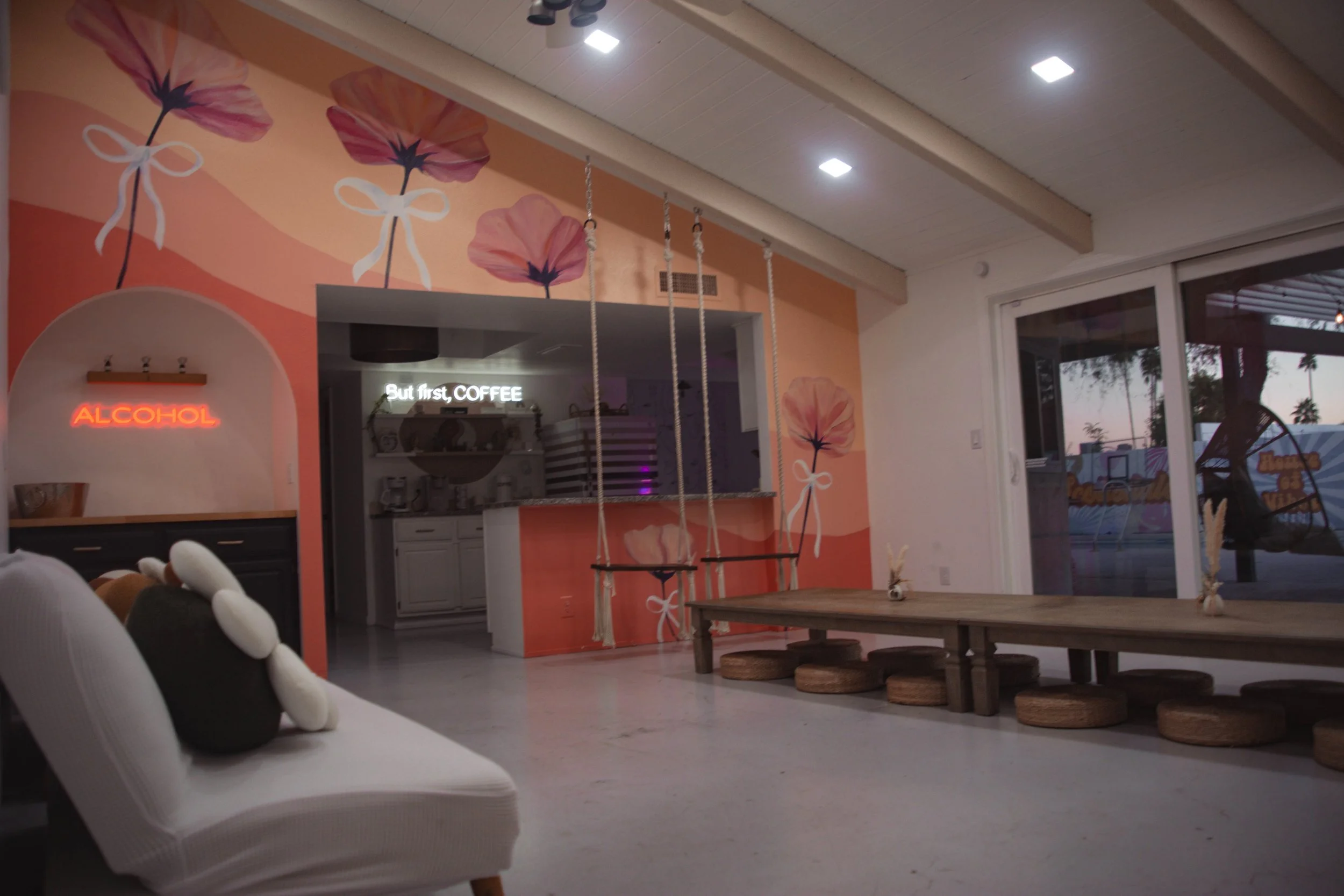 Indoor space with pink floral wall art, hanging swings, low wooden table with round cushions underneath, and a neon sign reading 'ALCOHOL' near a white couch with plush toys. Large window shows outdoor trees at dusk.