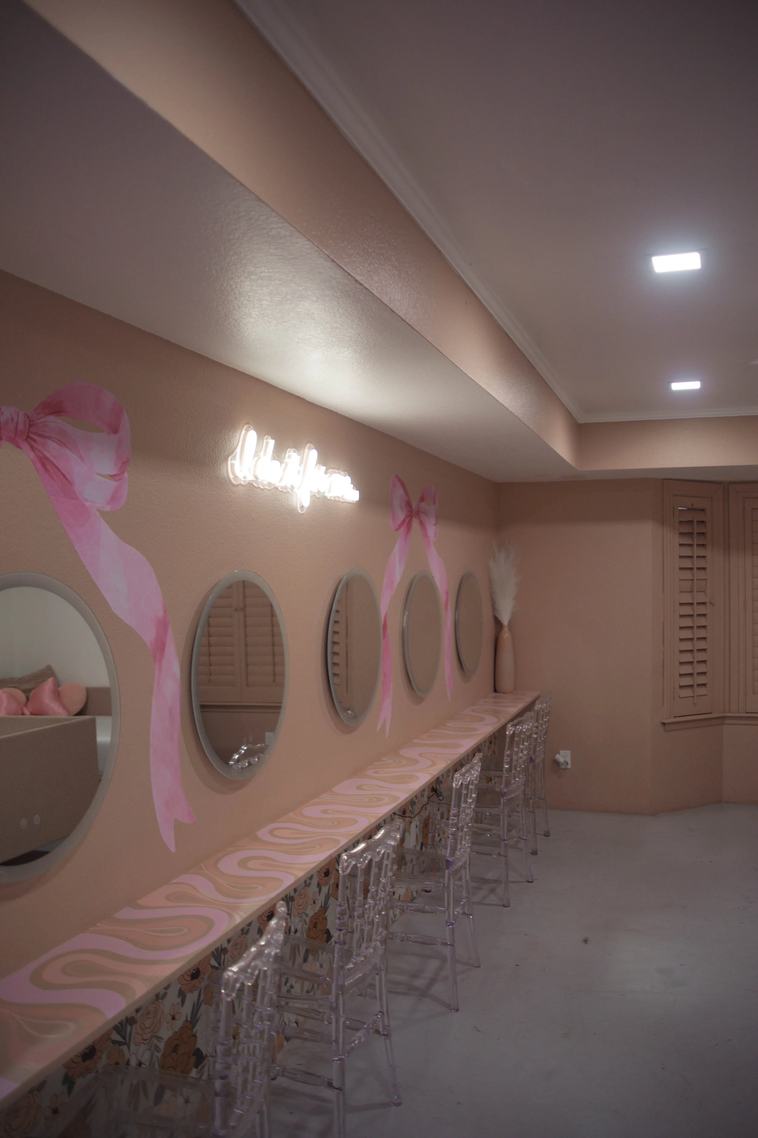 A decorated interior wall with pink ribbons, circular mirrors, and a neon sign that appears to say 'hello pink' in a pastel-colored room with transparent chairs and a vase with white pampas grass.