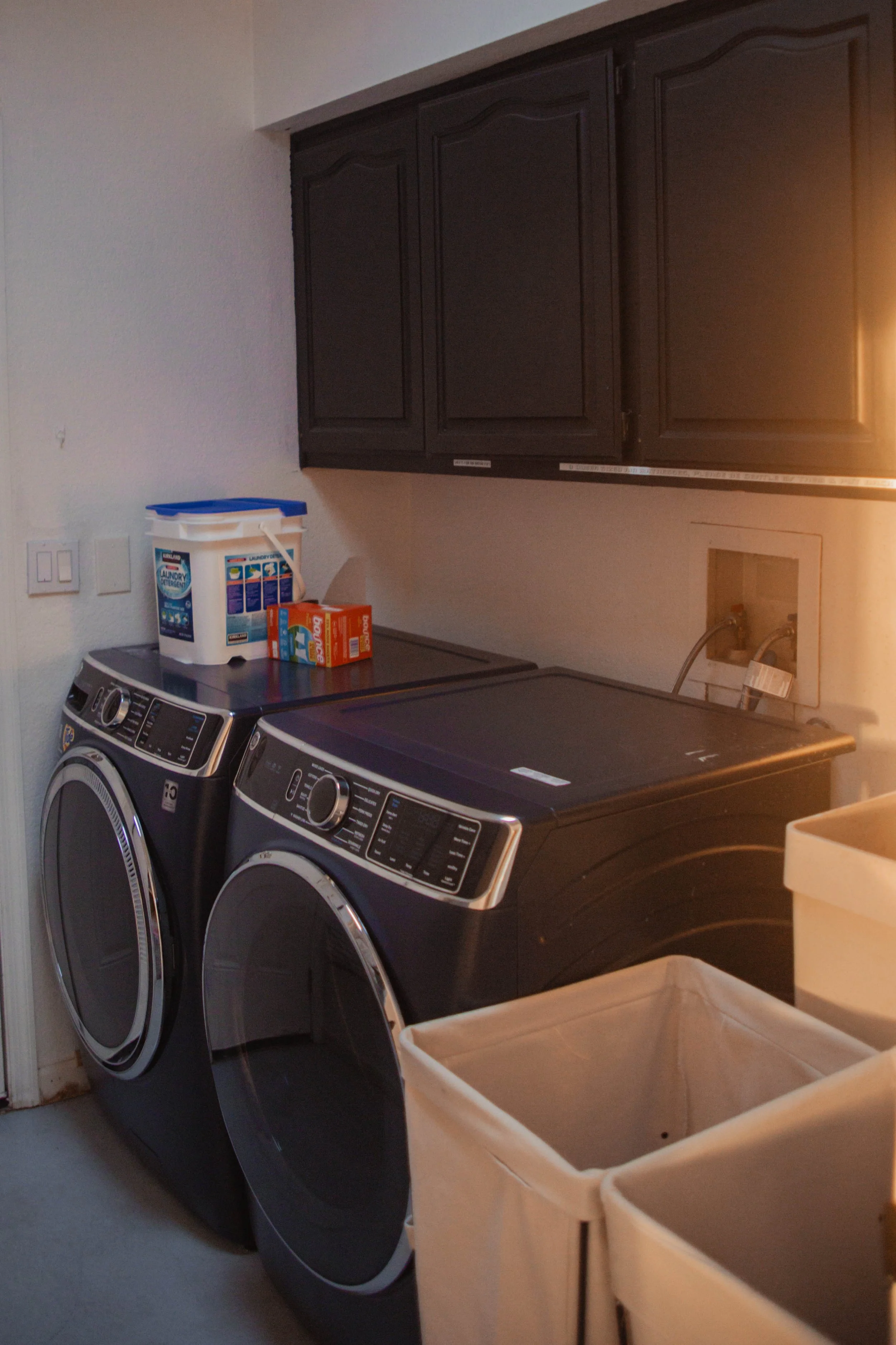 A laundry room with a dark-colored washer and dryer, a wall-mounted cabinet, laundry detergent, and fabric softener boxes on top of the appliances.