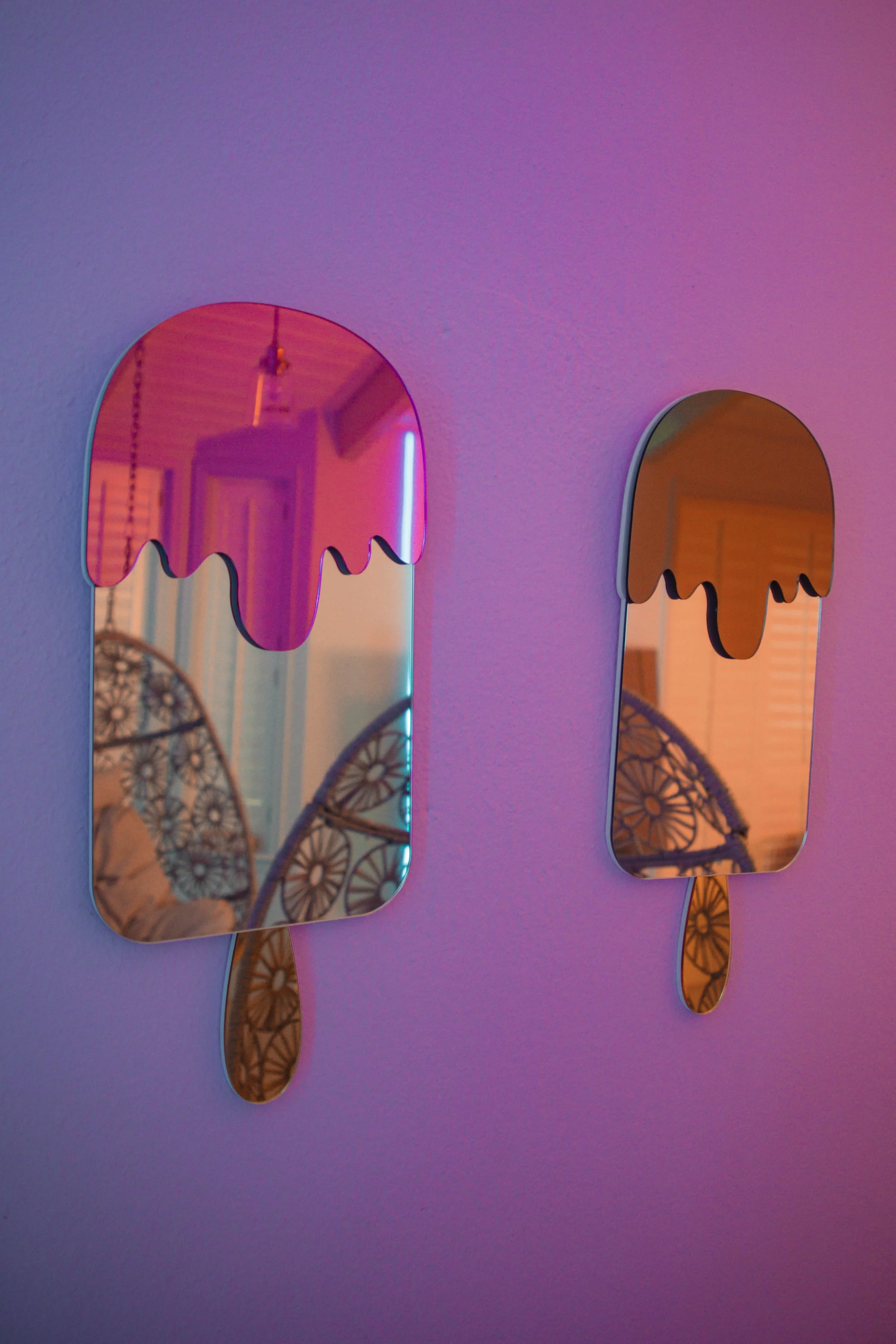 Two mirrors designed as melting ice cream popsicles with wooden stick handles, hanging on a wall, reflecting a room with wicker chairs and soft multicolored lighting.