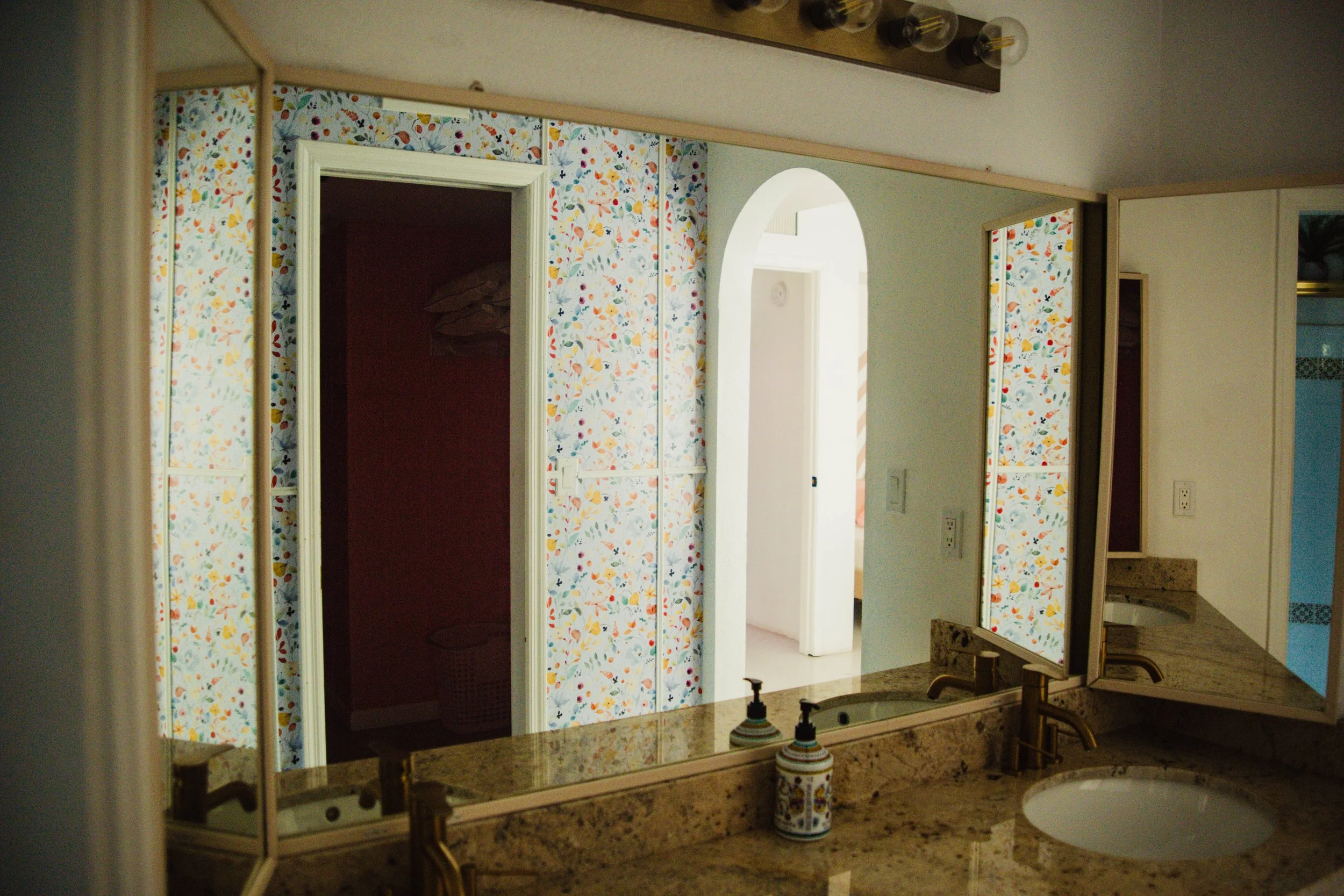 Bathroom mirror reflecting a speckled beige granite countertop and two bottles of hand soap. Two metal sinks with gold fixtures. Behind the mirror, a wall with floral wallpaper and a doorway leading to a dark laundry or closet space, with a laundry b