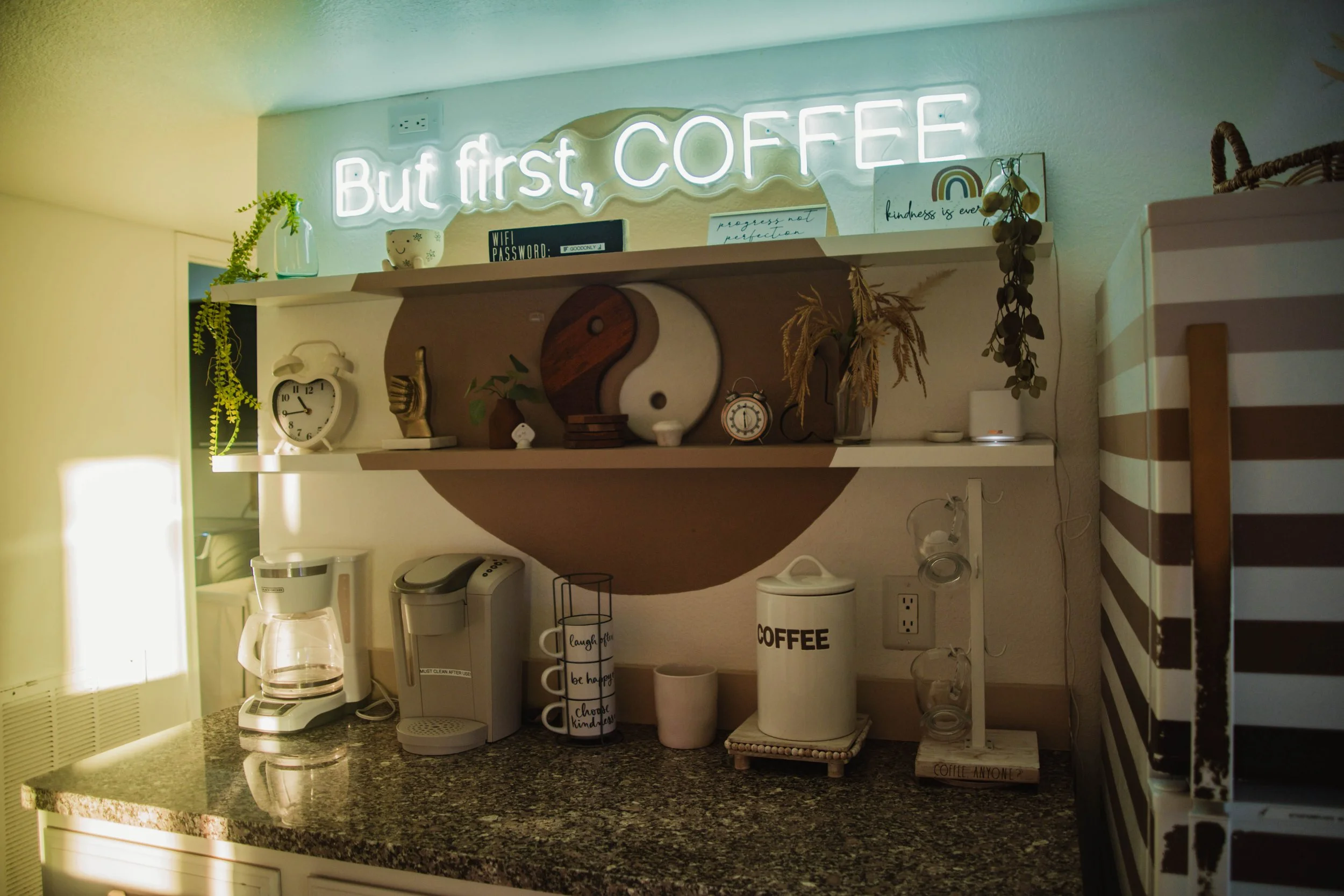 A cozy kitchen corner with a granite countertop holding a coffee maker, a kettle, and a coffee jar. Two shelves above hold decorative items, including a yin-yang symbol, plants, and signs that read 'But first, COFFEE' and motivational quotes. A neon 