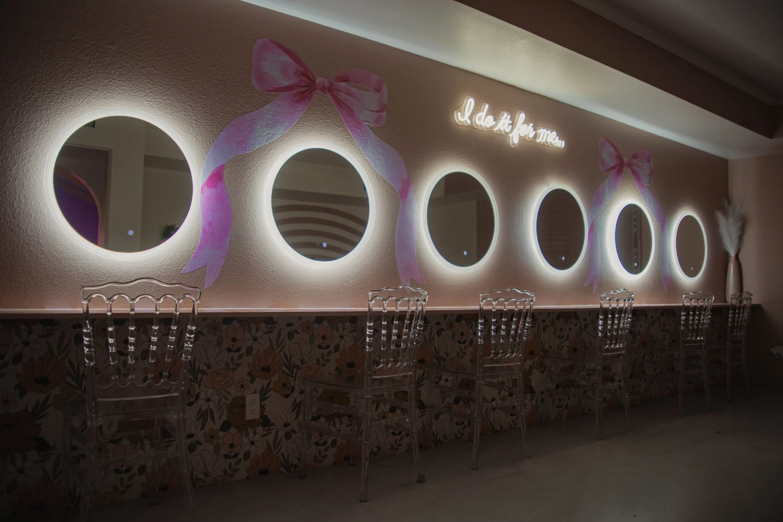 Decorative vanity area with five circular mirrors with backlit frames, pink bows, a neon sign saying 'I do it for me,' and transparent chairs arranged in front.