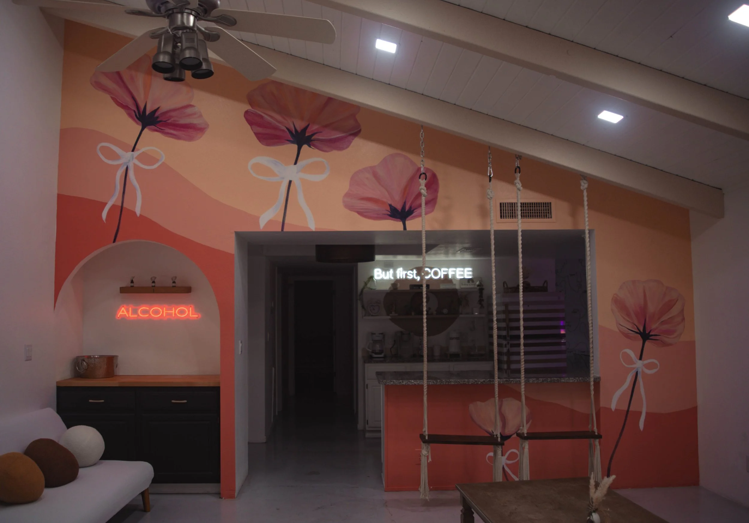 Interior of a cafe with a peach and pink floral mural on the wall, three large pink flowers with white ribbons, a ceiling fan, neon signs that read 'ALCOHOL' and 'But first, COFFEE', and a wooden swing with ropes.