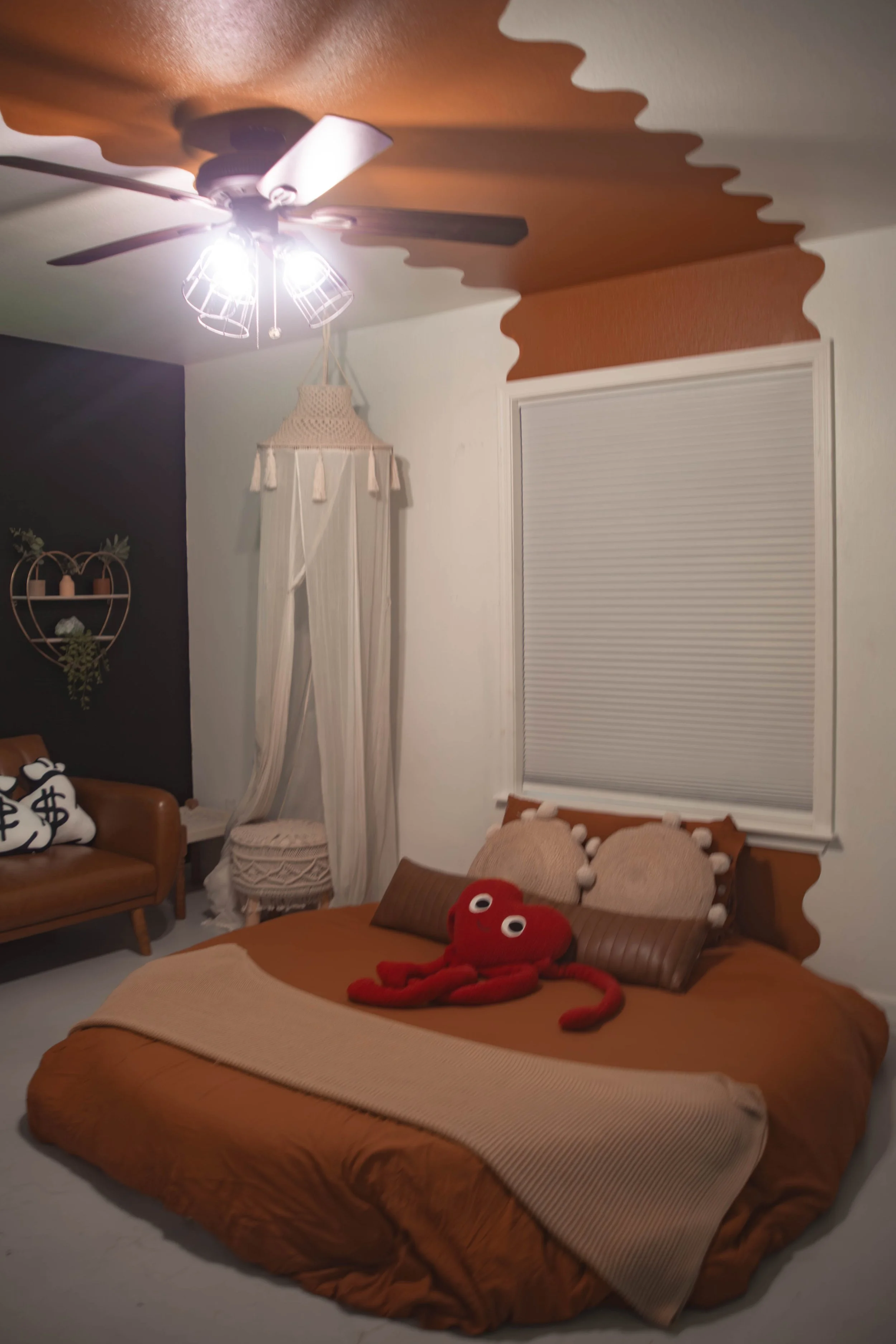 A bedroom with a brown bed, decorative pillows, and a red plush octopus toy on the bed. There is a window with closed blinds, a small wicker chandelier, and a white curtain with tassels. To the left, there is a brown leather sofa with plush pillows, 
