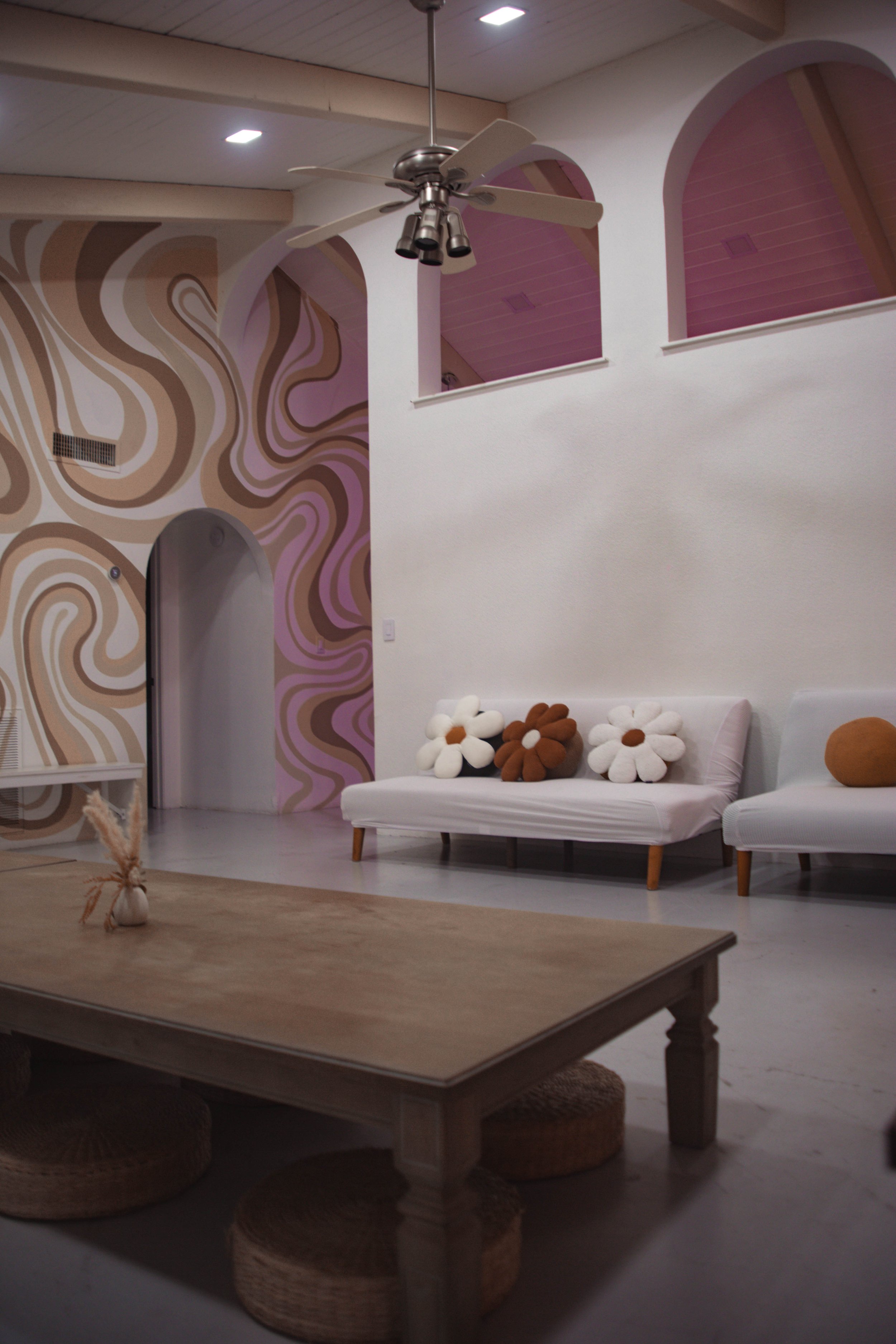 A cozy living room with beige and pink abstract wall art, a white couch with flower-shaped cushions, a wooden table with woven cushions underneath, and a ceiling fan in a bright, modern space.