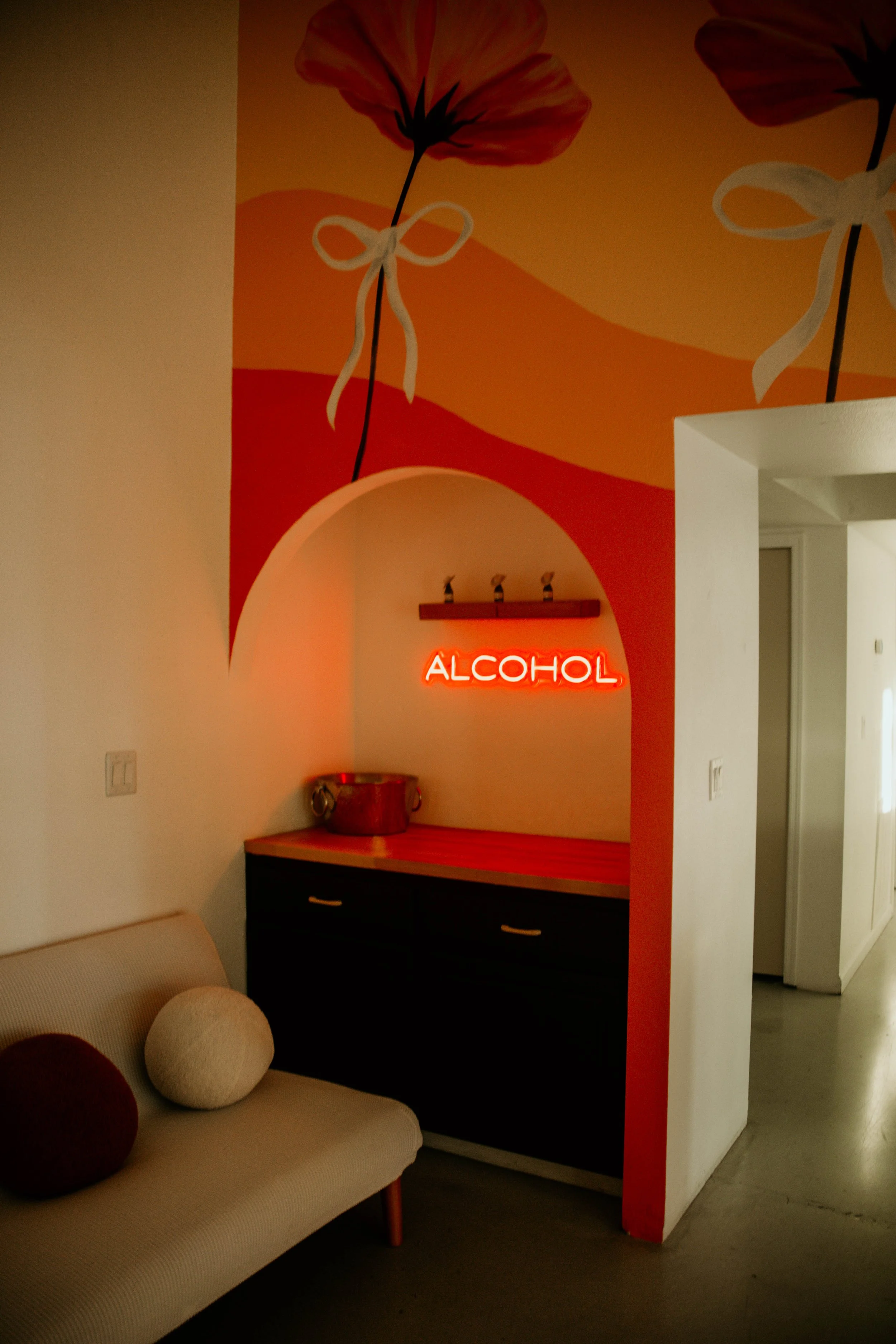 Interior decor with a wall featuring floral mural and a neon sign that says 'ALCOHOL' in red, a sideboard, and a beige sofa with three round pillows.