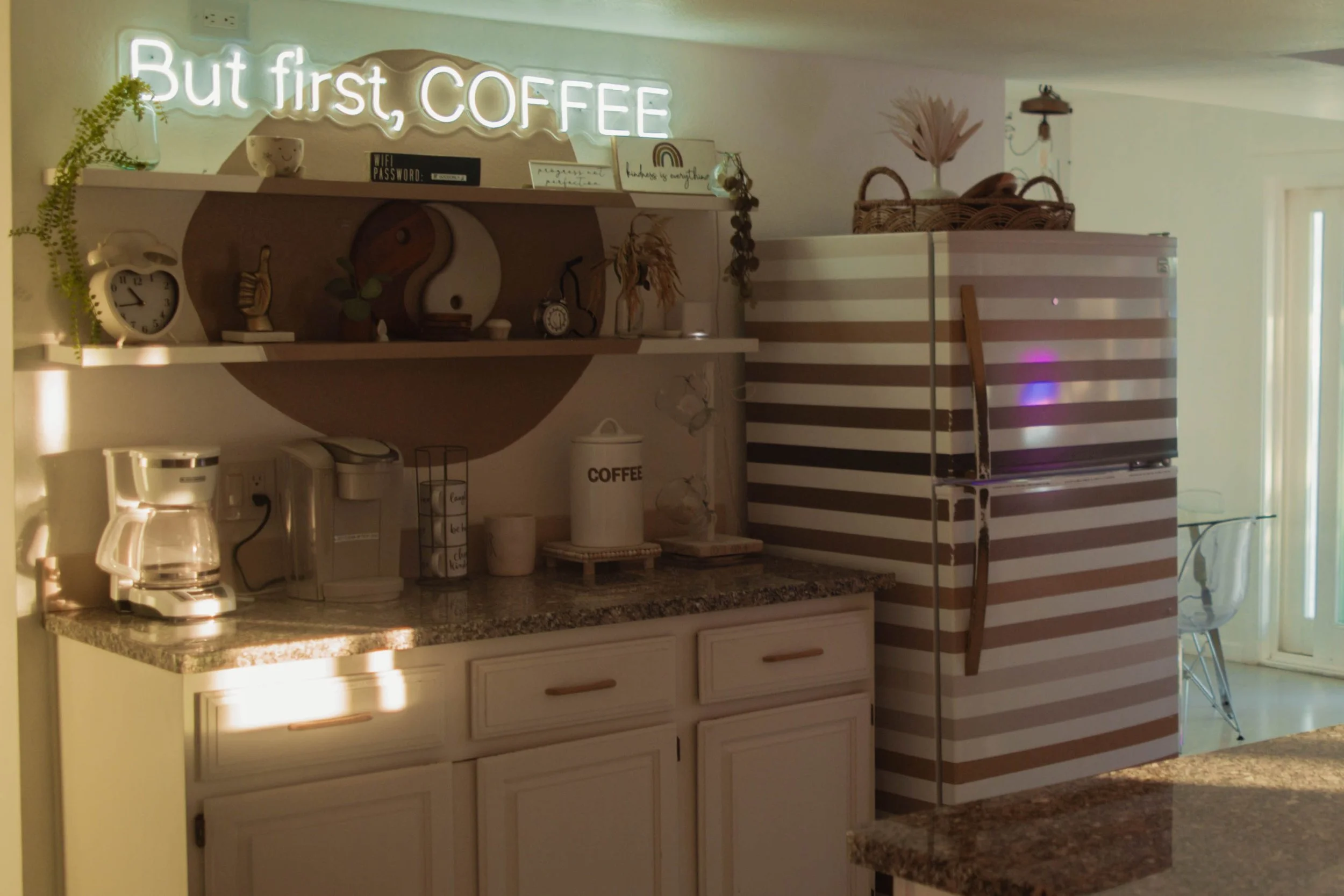 A kitchen corner with a striped refrigerator, granite countertops, and white cabinets; a backsplash decorated with artwork and plants; a coffee maker and toaster on the counter; a neon sign that says 'But first, COFFEE' above the shelf.