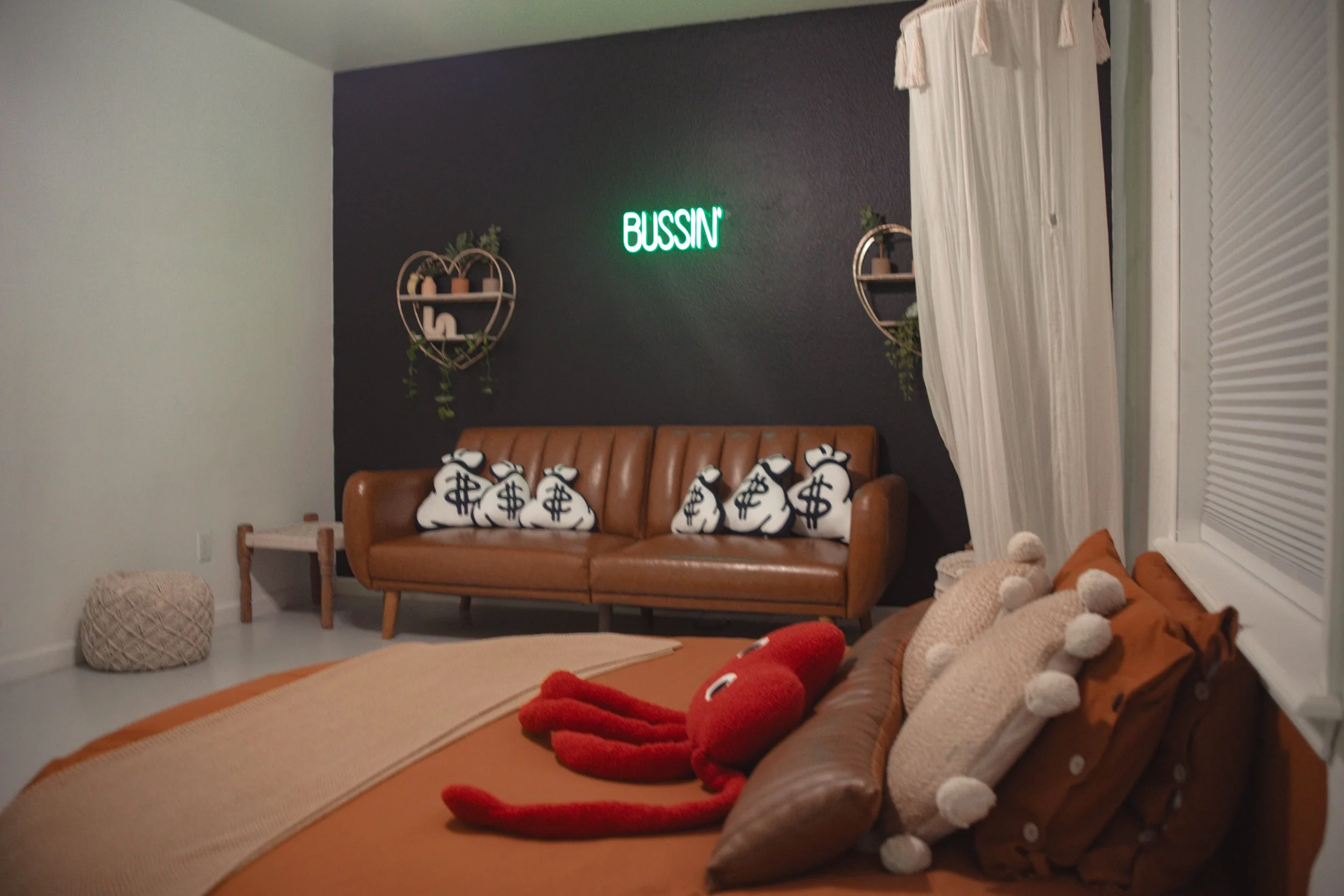 A cozy living room with a brown leather sofa adorned with pillows, including a red plush octopus, and a beige blanket. Behind the sofa, there is a black accent wall with a neon green sign reading 'BUSSIN' and two circular wall shelves with potted pla